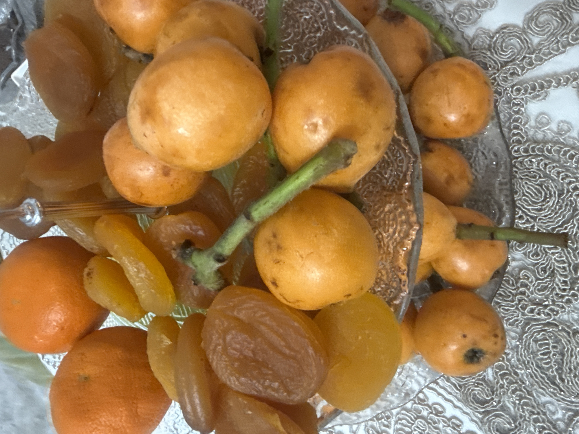 The image shows a variety of fruits on a glass plate. There are several loquats, some with stems still attached. Also present are oranges and dried apricots. The fruits are arranged on a patterned surface, possibly a tablecloth. The location is unknown.