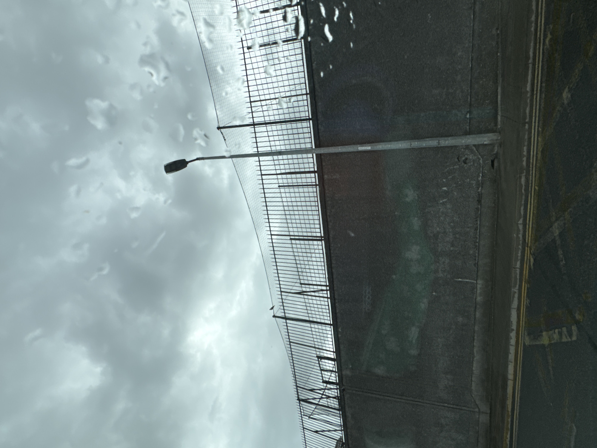 The image depicts a cloudy sky with rain droplets on the lens, obscuring the view of a grey concrete wall topped with a black metal fence. A tall, grey lamppost stands in front of the fence. In the distance, a single bird is perched on the fence. The wall itself appears to have some faint, light-colored markings or graffiti on its surface. In the foreground, a dark asphalt road with double yellow lines is visible. The scene suggests an overcast day in an urban environment. The location is identified as Waterford, Ireland.