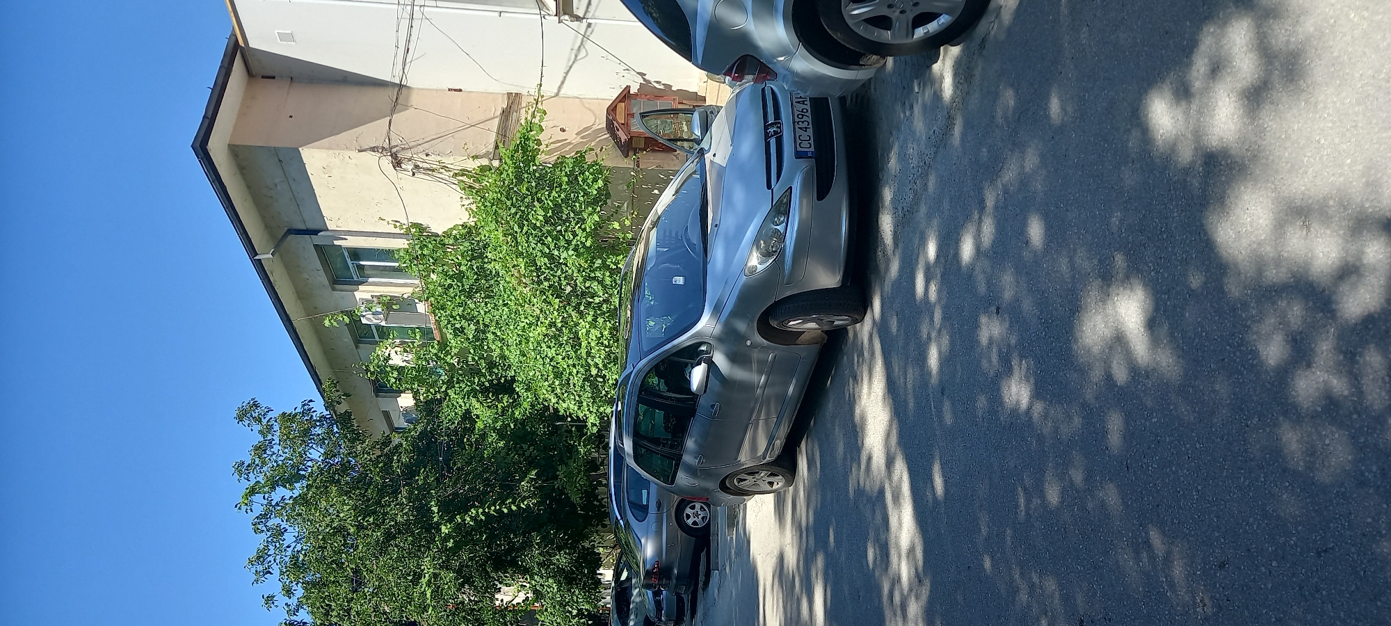 In the city of Silistra, Bulgaria, a silver Peugeot 307 wagon is parked on an asphalt surface, partially shaded by trees. The car's front passenger door is open. The license plate reads "CC 4396 AH". Another silver car is parked next to it. In the background, a light-colored building with several windows and an air conditioning unit is visible. Lush green trees and vegetation surround the building and cars. The sky is clear and blue.