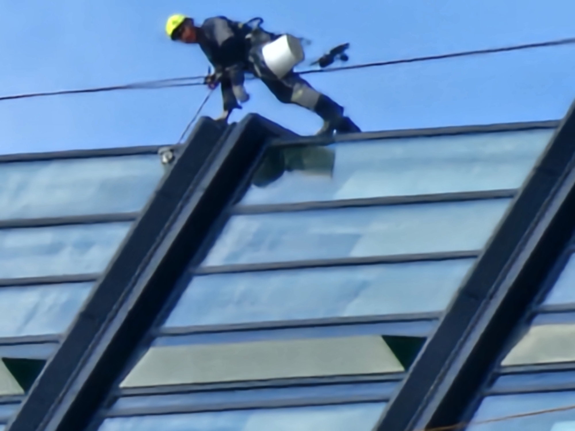 The image shows a window cleaner at work on the exterior of a building with a glass facade. The worker is wearing a yellow hard hat and a gray uniform, with a harness and safety lines. A white bucket is attached to their waist. The worker is positioned on the edge of the roof, leaning forward and appears to be in the process of cleaning the windows. The building's exterior consists of large, reflective glass panels framed by dark structural elements.
The scene is set against a clear blue sky, suggesting a daytime setting with fair weather. The angle of the shot indicates the photo was taken from below, looking up at the worker. The reflective glass panels show hints of the surrounding environment, possibly other buildings or sky. There are no visible text elements in the image. The overall impression is one of a high-rise worker engaged in a potentially hazardous but routine task. Given the prompt's context, this scene is situated in Moscow, Russia.
