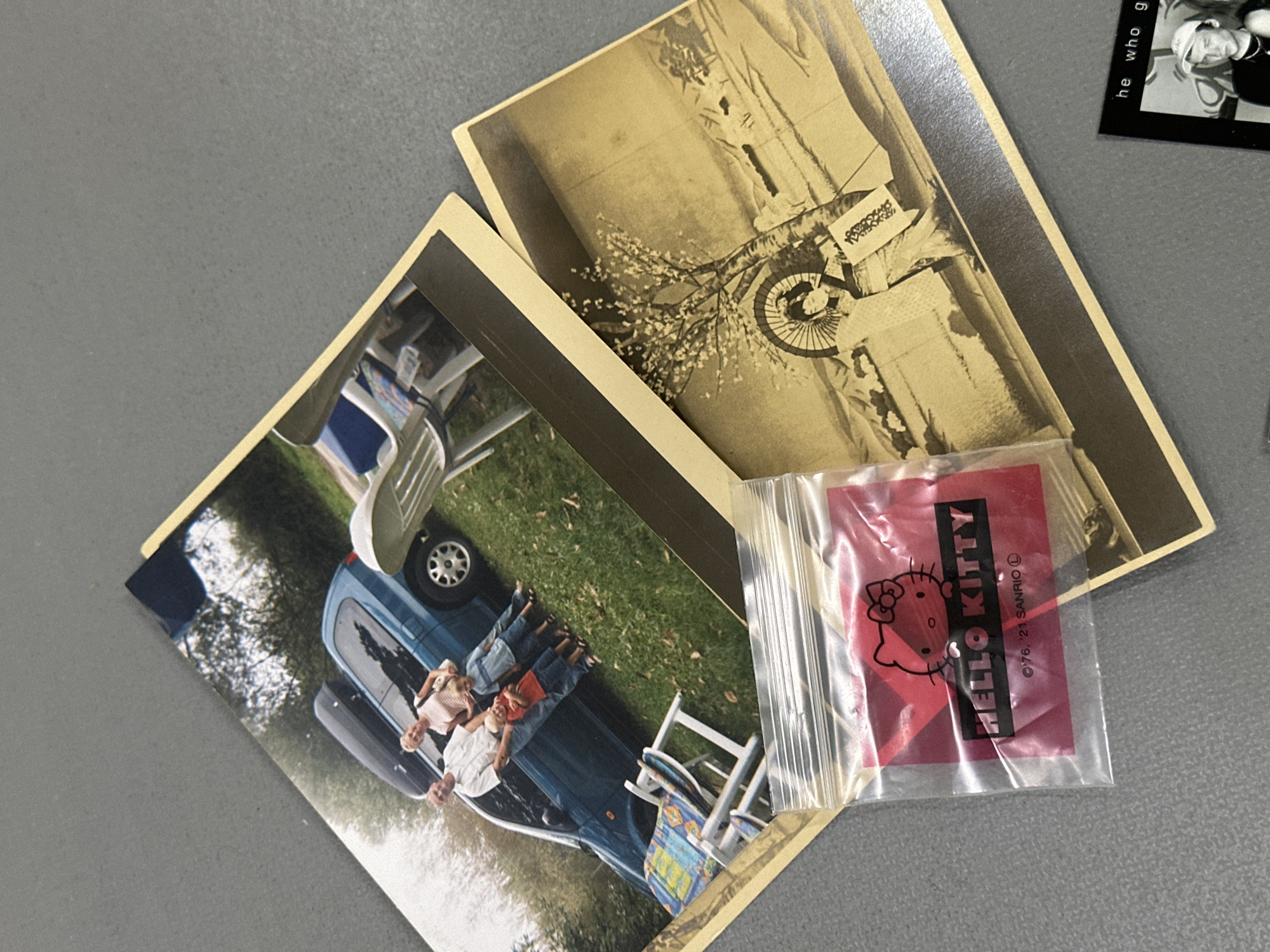 The scene shows three photos on a grey surface.
One photo features a blue vehicle parked on grass with several individuals lying on its hood. Nearby are two white folding chairs.
A second photo is a sepia-toned image depicting a person on a bicycle in front of a building, which appears to be in Tunbridge Wells based on the architectural style and street sign.
A small, clear plastic bag containing a red rectangle with a Hello Kitty design is positioned on top of the photos.
A partially visible black and white photograph can be seen at the top right edge.