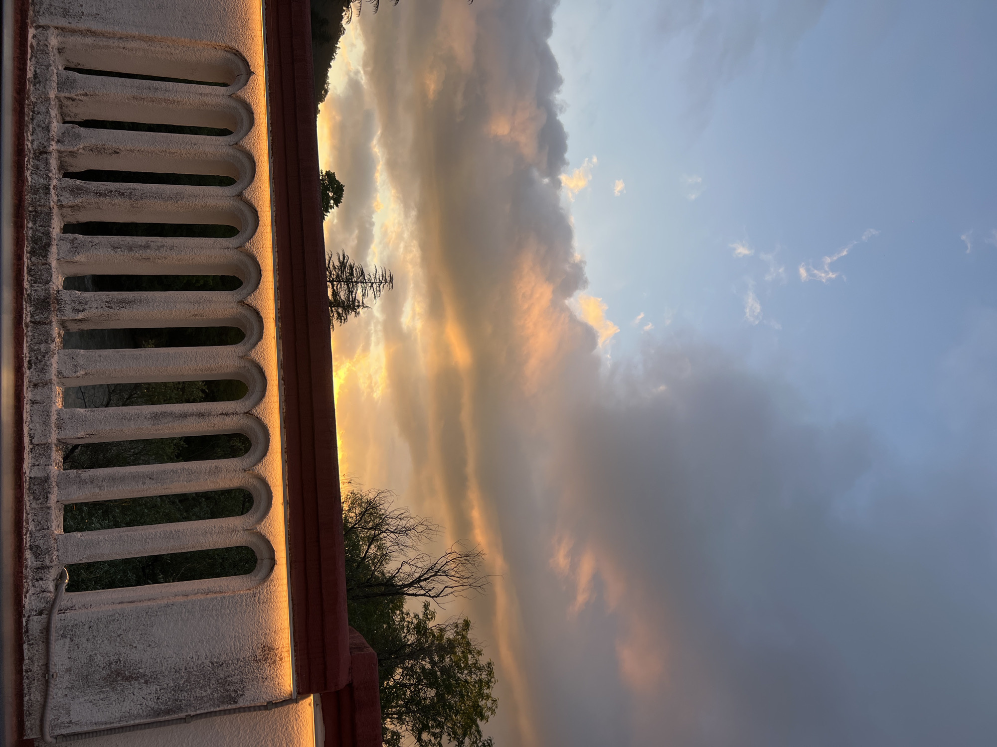 An outdoor, low-angle, full shot captures a balustrade with arched openings in the foreground, illuminated by a strip of warm light beneath a red roof. Beyond the balustrade, dark trees and a hillside are visible. The sky above displays a dramatic sunset with orange and pink clouds against a gradient of blue. The image is set in an unspecified location within an unknown city.