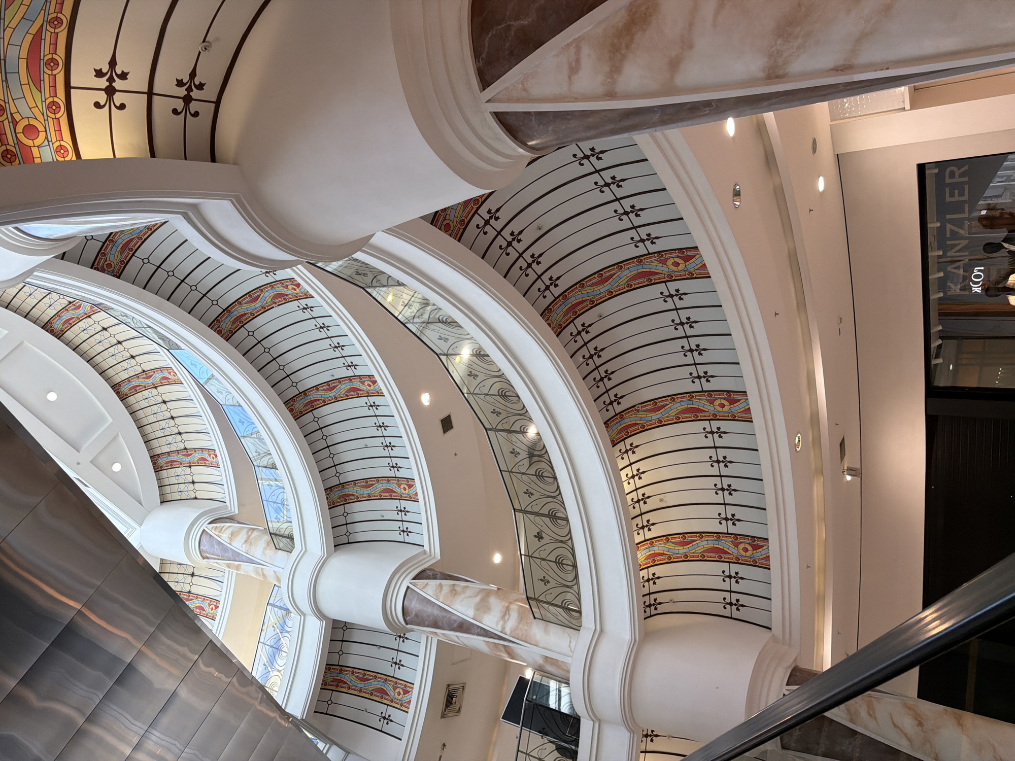 This image shows an interior view of a building, potentially a shopping center, characterized by elaborate architectural details. The architecture features rounded arches, decorative columns, and patterned ceilings with linear and floral motifs in earth tones and white. The scene is captured from a low angle, looking up towards the ornate ceilings and structural elements. There is an escalator visible on the left side of the image. The view is from the location of Unknown, Unknown.