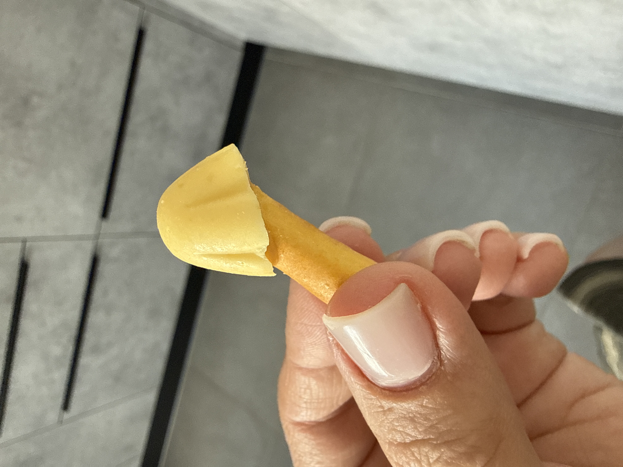 A close-up shot captures a hand holding a snack. The snack appears to be a cheese puff or a similar extruded snack, with a yellow, mushroom-cap-shaped piece of cheese or a similar topping adorning its top. The hand has neatly manicured nails, painted a light, glossy pink. The background is out of focus, showing a textured grey surface, possibly a kitchen cabinet or countertop, with dark horizontal lines. The image does not provide any information about the city of Lyubertsy, Russia, or any specific urban environment.