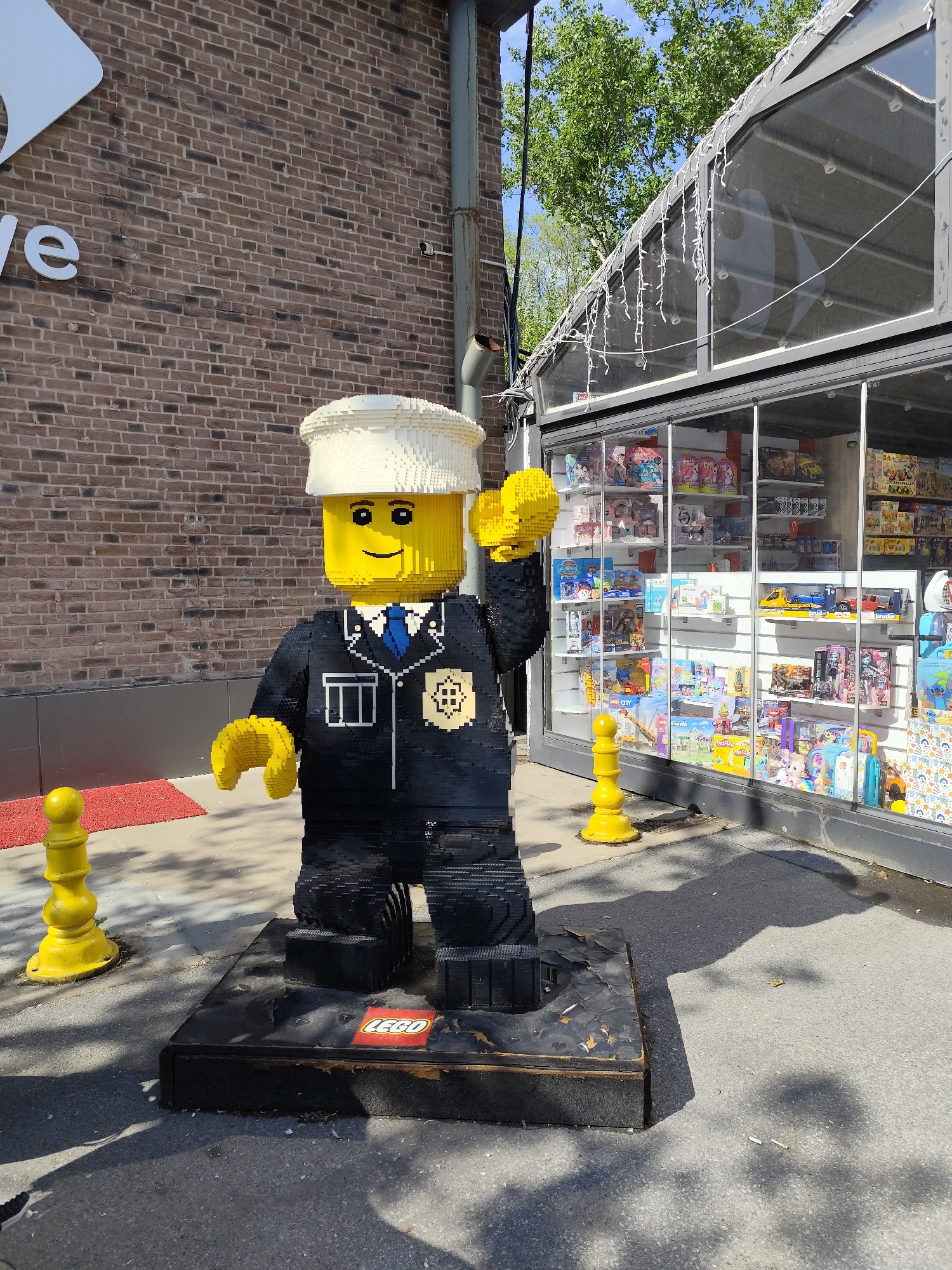 A life-sized LEGO sculpture of a police officer stands on a black platform labeled "LEGO" in front of a store. The officer figure is made of LEGO bricks, features a smiling face, a white cap, and a black uniform with a blue tie and badge. The sculpture is surrounded by yellow bollards. The background includes a brick building with the letters "ve" visible and a store with glass windows displaying various toys and merchandise. The environment suggests an outdoor city setting during the day.