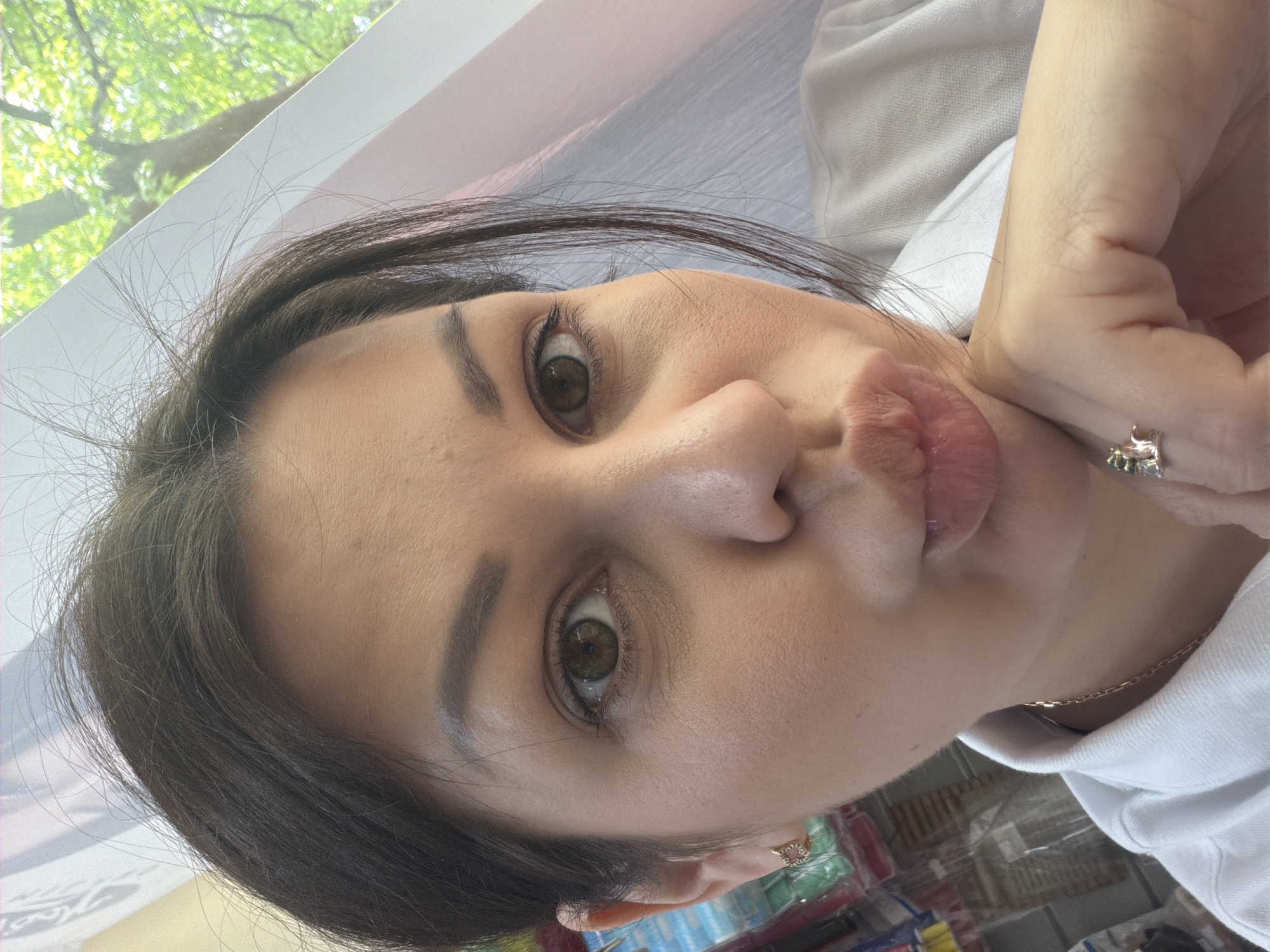 The image shows a close-up, selfie-style shot of a woman in Sochi, Russia. She has brown hair and brown eyes and is wearing a white collared shirt and gold jewelry, including earrings and a necklace. Her face is angled towards the camera, with her chin resting on her hand, and her lips are pursed. The background is blurry and appears to be an indoor setting with some foliage visible through what might be a window or screen. The overall impression is of a casual, personal moment captured outdoors.