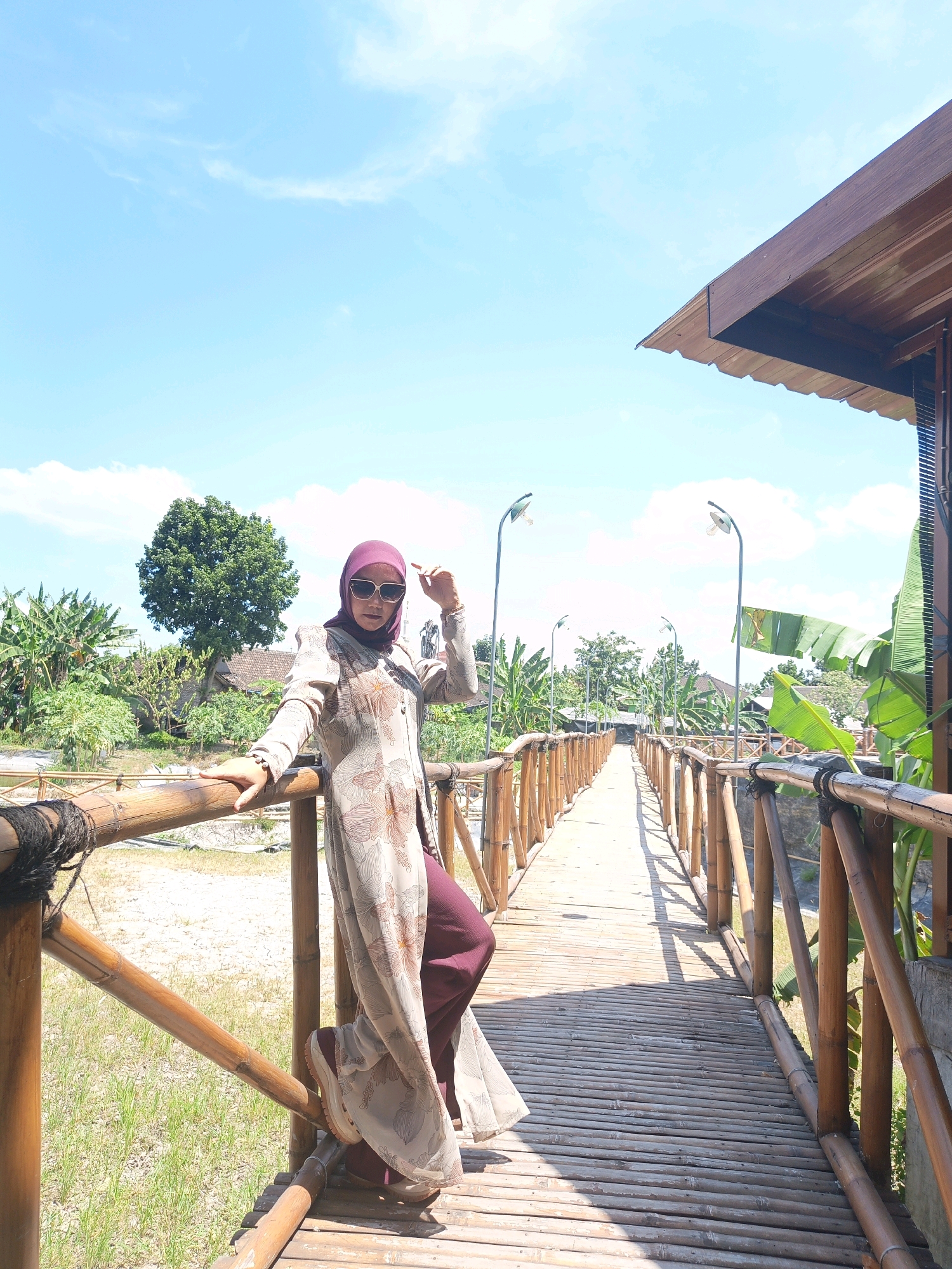A woman wearing a hijab and sunglasses poses on a bamboo bridge in Jonggrangan, Indonesia. She is wearing a long, light-colored dress with a floral pattern and maroon pants. The bridge, made of bamboo, stretches into the distance, lined with bamboo railings and posts. The background features lush greenery, including banana trees and a large tree, with a clear blue sky overhead. A part of a wooden structure with a brown roof is visible on the right side of the image. Streetlights are mounted along the bridge's railing. The woman is leaning on the railing with her left hand and has her right hand near her head. The scene appears to be outdoors on a sunny day.