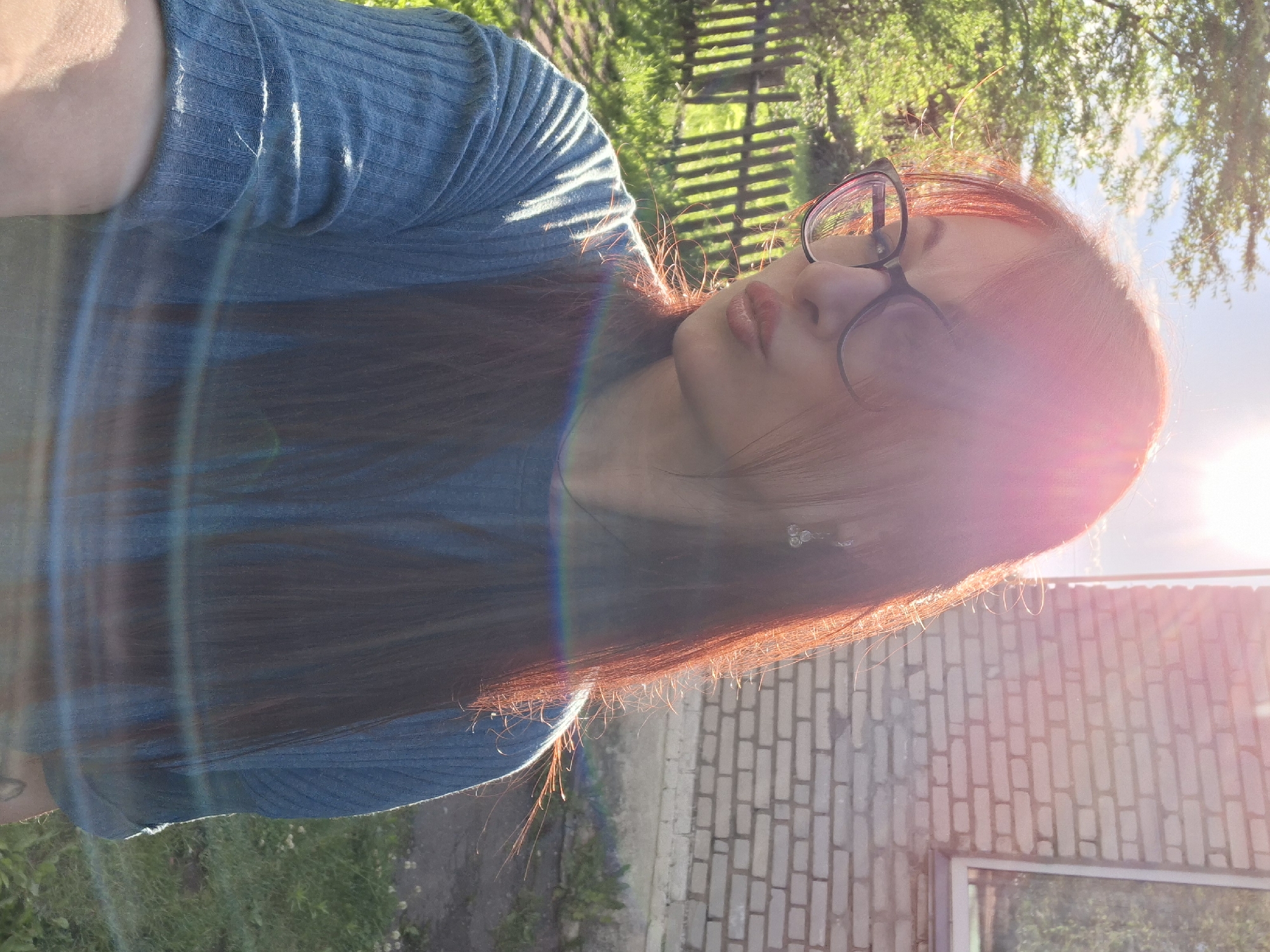 The image captures a selfie of a woman with long, reddish-brown hair, wearing black-rimmed glasses and a blue, ribbed long-sleeved shirt. She is outdoors, with a wooden fence and greenery visible behind her, suggesting a rural or suburban setting. A brick building with a window is to her right. The sun is low in the sky, casting a warm, bright light that creates lens flare and highlights her hair. She is puckering her lips, looking to her left. The scene appears to be in the Забалоцкі сельскі Савет region of Belarus, given the context of the image's origin.
