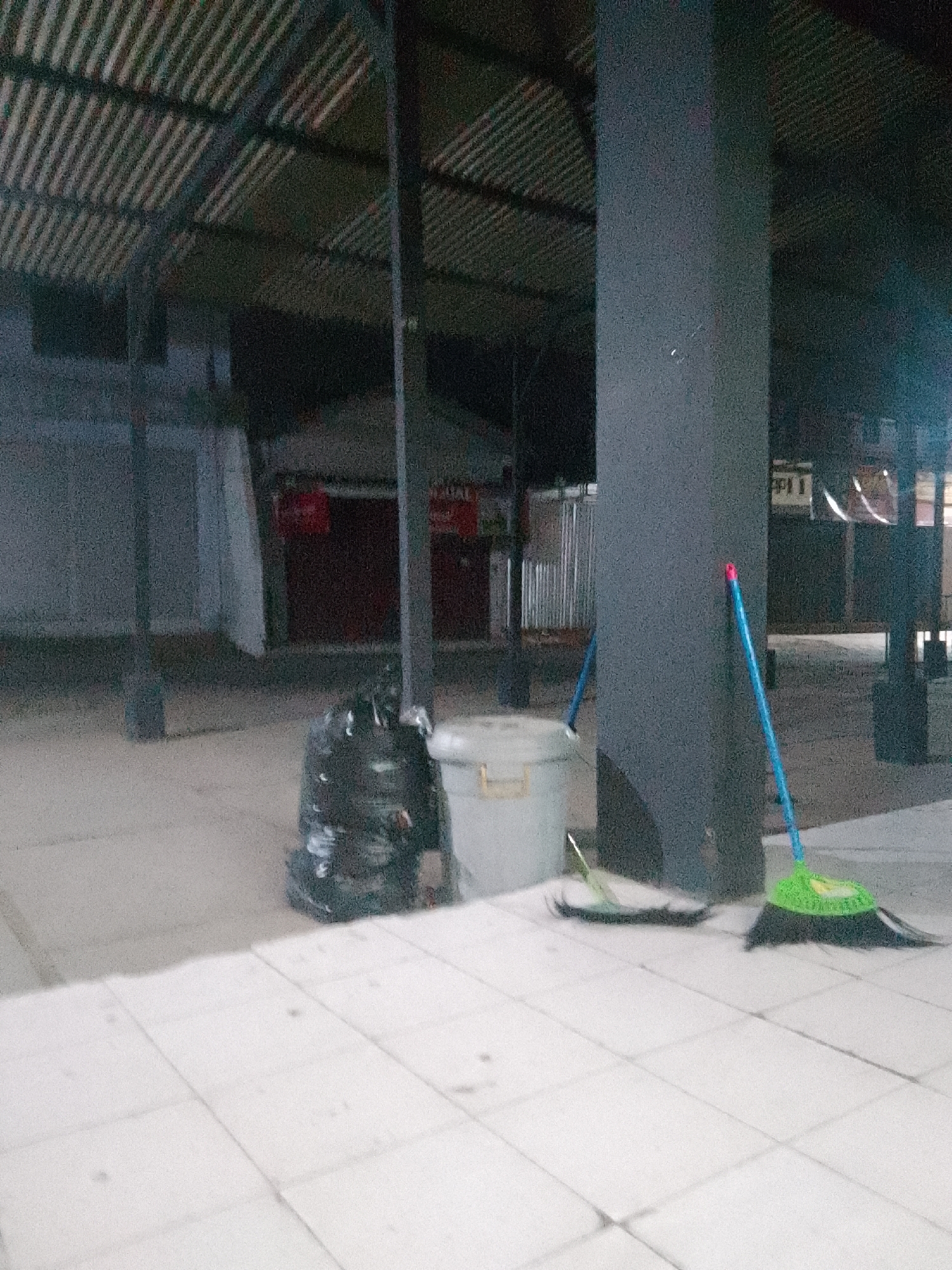The image depicts an outdoor scene at what appears to be a covered market or a public waiting area in Trenggalek, Indonesia. The environment is characterized by a tiled floor and a corrugated metal roof supported by pillars. In the foreground, a large black trash bag is placed on the ground next to a light-colored plastic bin. Two brooms are also visible: one with a green head and a blue handle, leaning against a dark gray pillar, and another with a lighter-colored head and handle lying on the ground to the left. In the background, there are structures that resemble market stalls with closed shutters, some of which are painted red. The lighting suggests it is either early morning, late evening, or an overcast day. There are no people visible in the frame.
