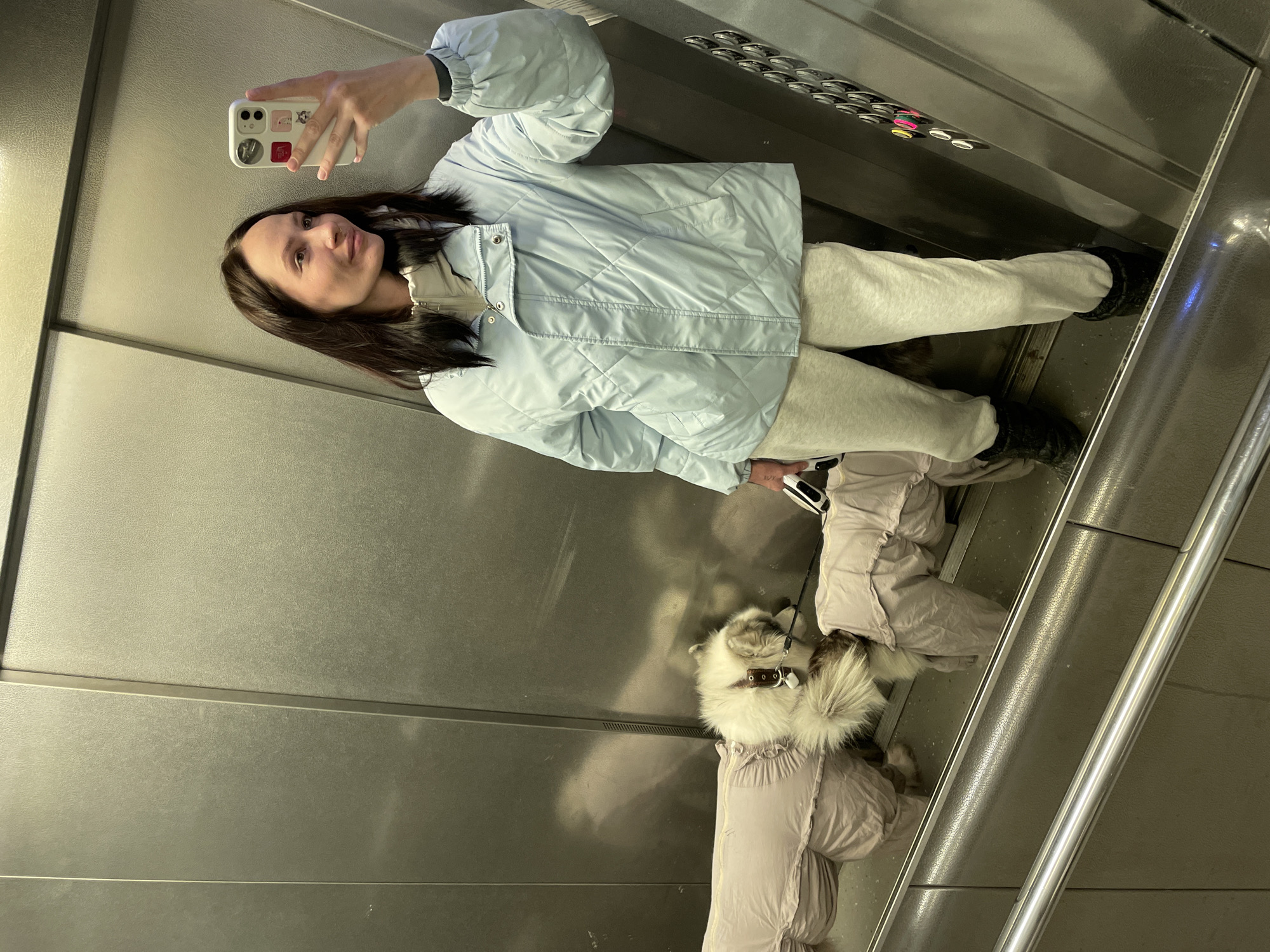 A woman and a small dog are inside an elevator with brushed aluminum or stainless steel walls. The woman is facing the elevator's mirrored wall and holding up her smartphone, presumably taking a selfie or video. She is wearing a light blue quilted jacket, light gray sweatpants, and black boots. The dog is wearing a lavender-colored coat with a faux fur collar. The elevator's control panel is visible in the background, featuring rows of buttons. The dog is leashed. The location may be in Izhevsk, Russia, but the image itself does not provide specific indicators of the city.