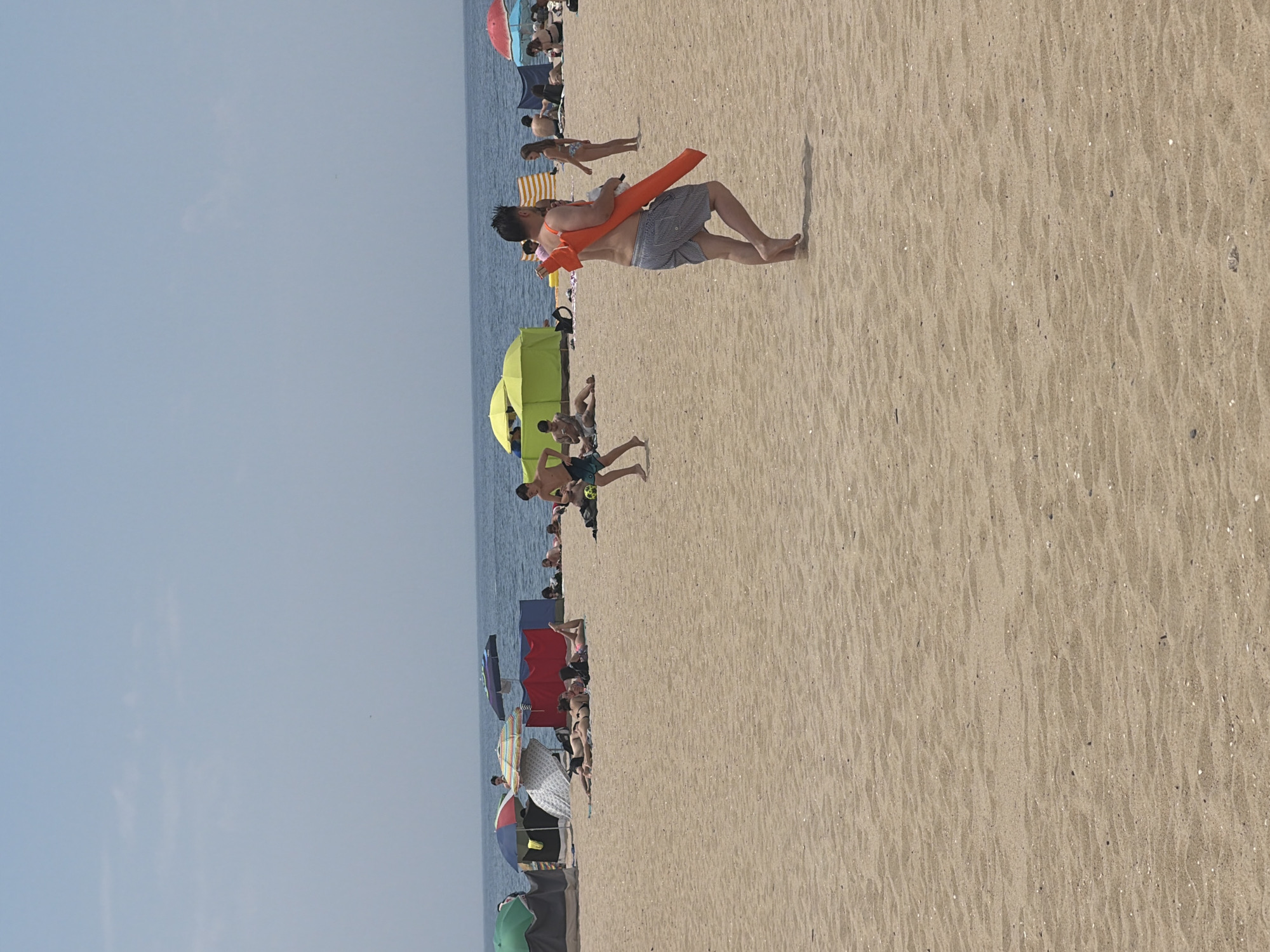The image shows a crowded beach scene on a sunny day. In the foreground, a man in blue and white striped shorts is walking with an orange float slung over his shoulder. He is facing away from the camera. Further back, several people are sunbathing or relaxing under beach umbrellas and tents. Some individuals are visible in the ocean. The beach is sandy, with the ocean in the background under a clear blue sky. This scene is typical of Espinho's Praia da Baía, known for its lively atmosphere during summer months.