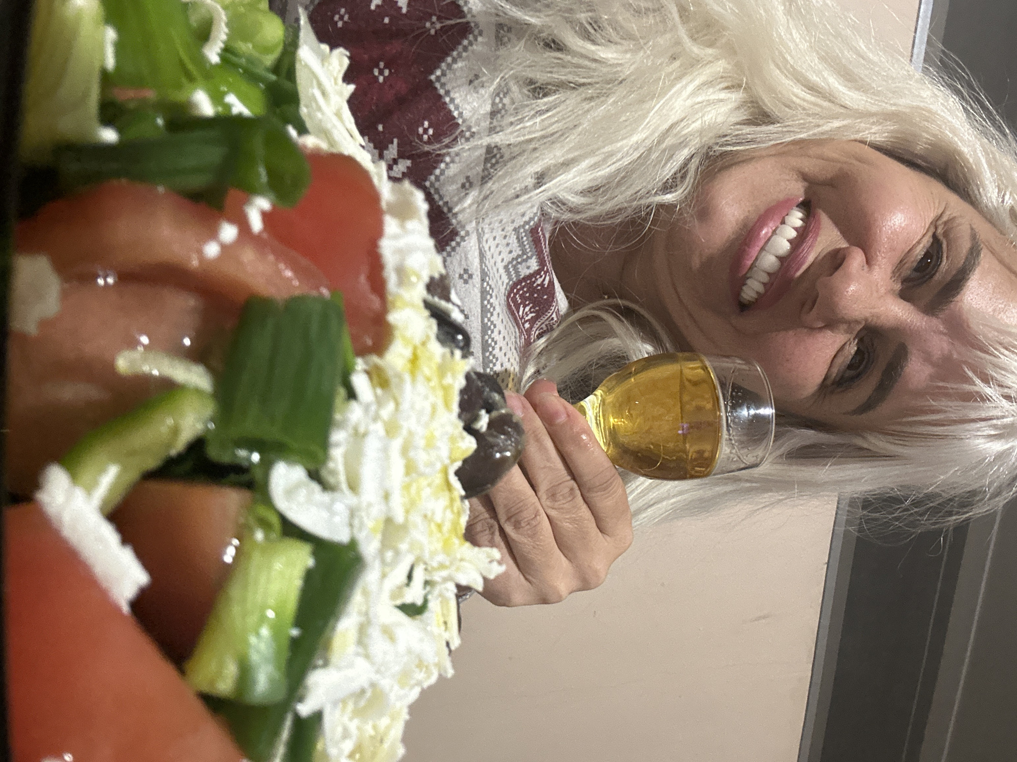 The image depicts a woman with blond hair holding a glass of amber-colored liquid. Next to her is a salad of tomatoes, cucumbers, green onions, feta cheese, and olives. She's wearing a sweater with a winter-themed pattern. The background suggests an indoor setting. The city and country aren't identifiable from visual cues in the image.