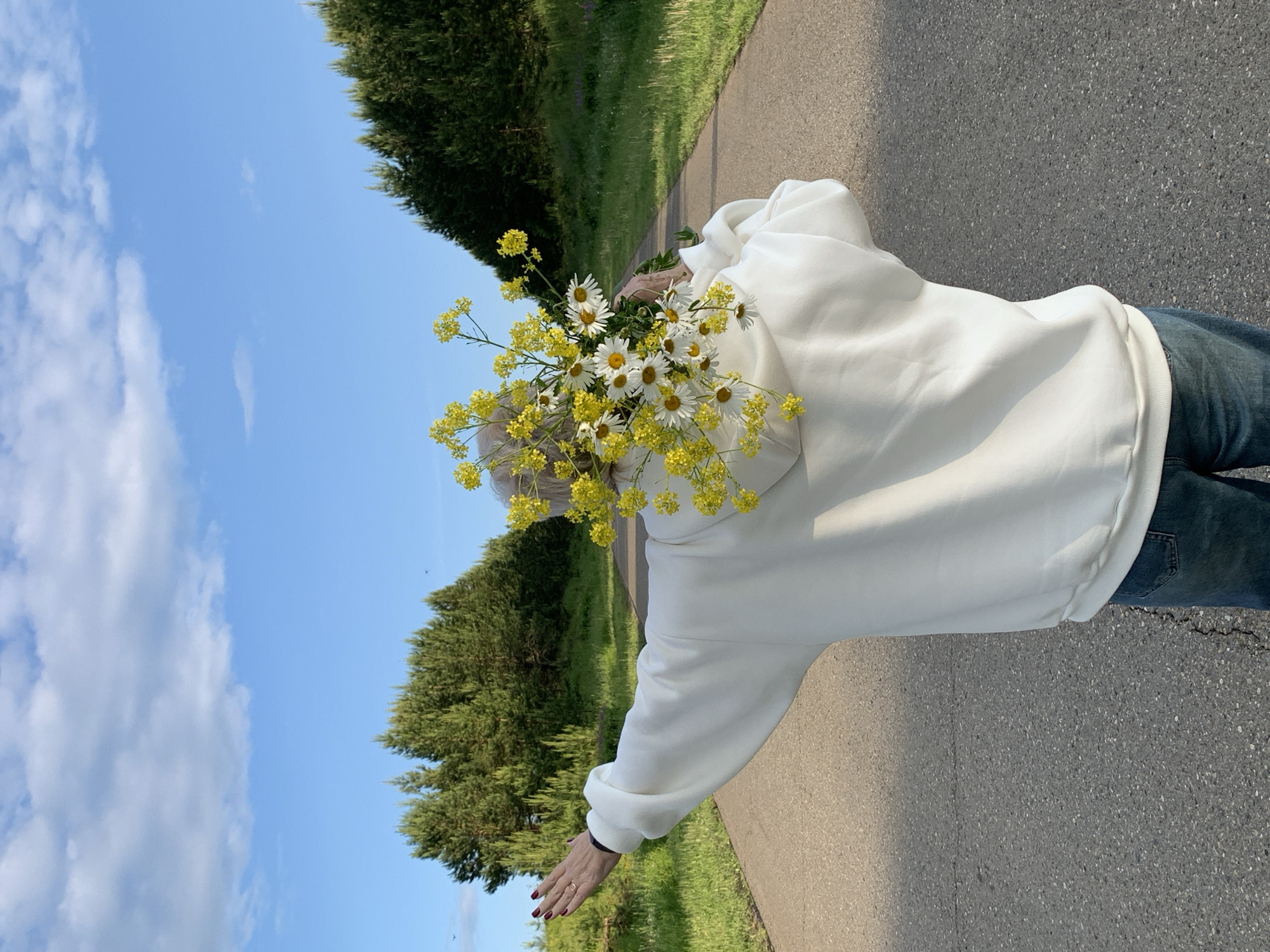 The image, when viewed upright, depicts a person outdoors, walking or standing on a paved path in what appears to be a rural or semi-rural setting in Bol'shoi Bukor, Russia. The person is seen from a back/side angle, wearing a white hooded sweatshirt and blue jeans. Their head is entirely obscured by a large, vibrant bouquet of flowers held in front of them. The bouquet is composed predominantly of white daisies with yellow centers, interspersed with numerous clusters of small bright yellow flowers, likely a type of wild mustard or rapeseed.
The person's right arm is extended forward, with their hand visible. The fingernails are painted a dark red, and a simple ring is worn on the ring finger. The background features a clear blue sky with some scattered white clouds on the left side. A dense line of green trees, appearing to be a mix of conifers and deciduous types, borders the right side of the path, with green grass visible between the trees and the path. The path itself is dark grey and textured. The scene is illuminated by bright daylight, suggesting a sunny day. No text is visible in the image.