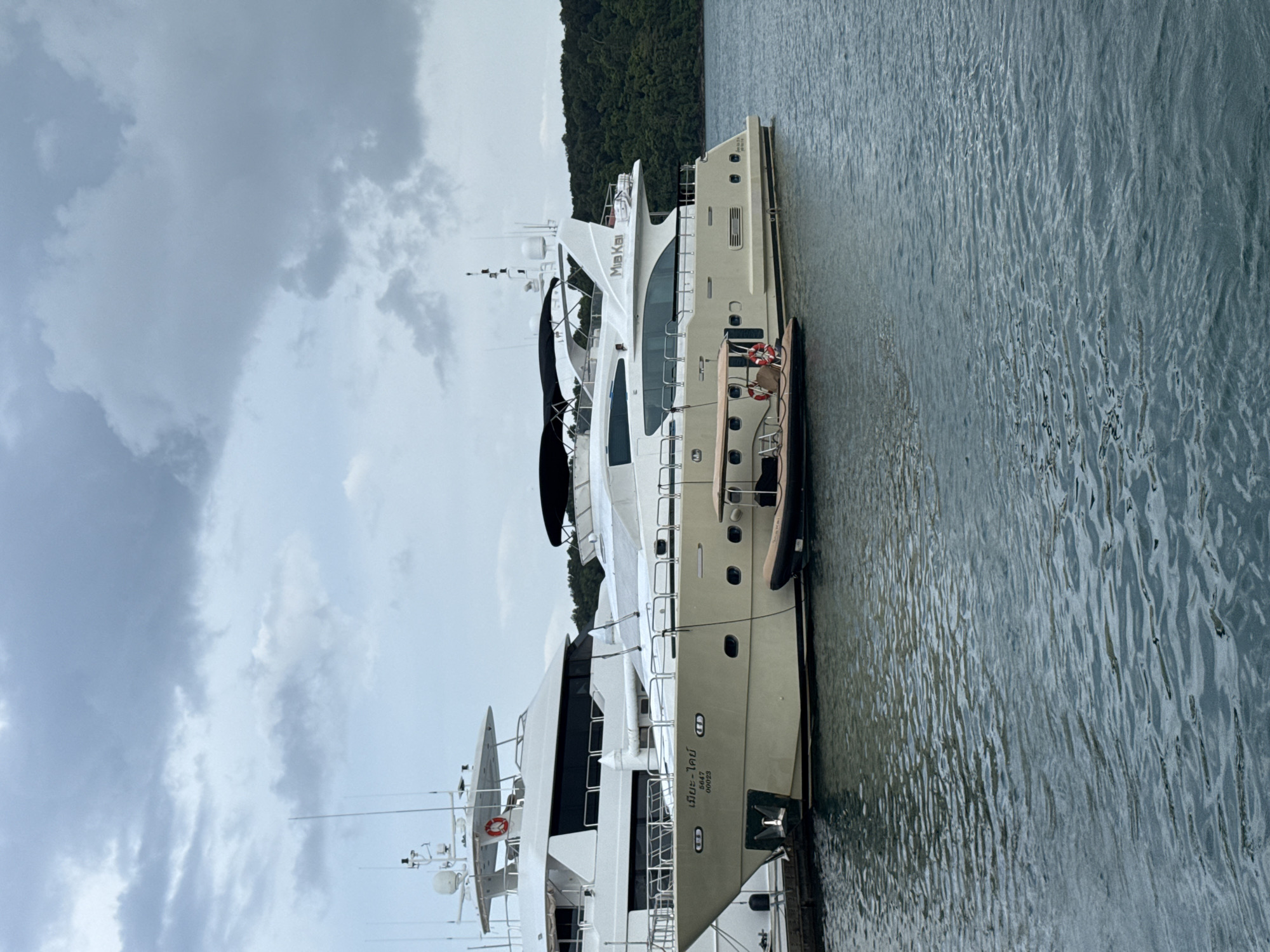 An anchored tender boat is positioned alongside a docked luxury yacht under a cloudy sky in the city of Pa Khlok, Thailand. The yacht, which is beige with a white superstructure, is moored at a dock. The tender boat is brown and has a life preserver attached to its side. In the background, a larger white yacht is docked to the left of the beige yacht. A forested hillside rises behind the yachts. The water is calm with ripples.