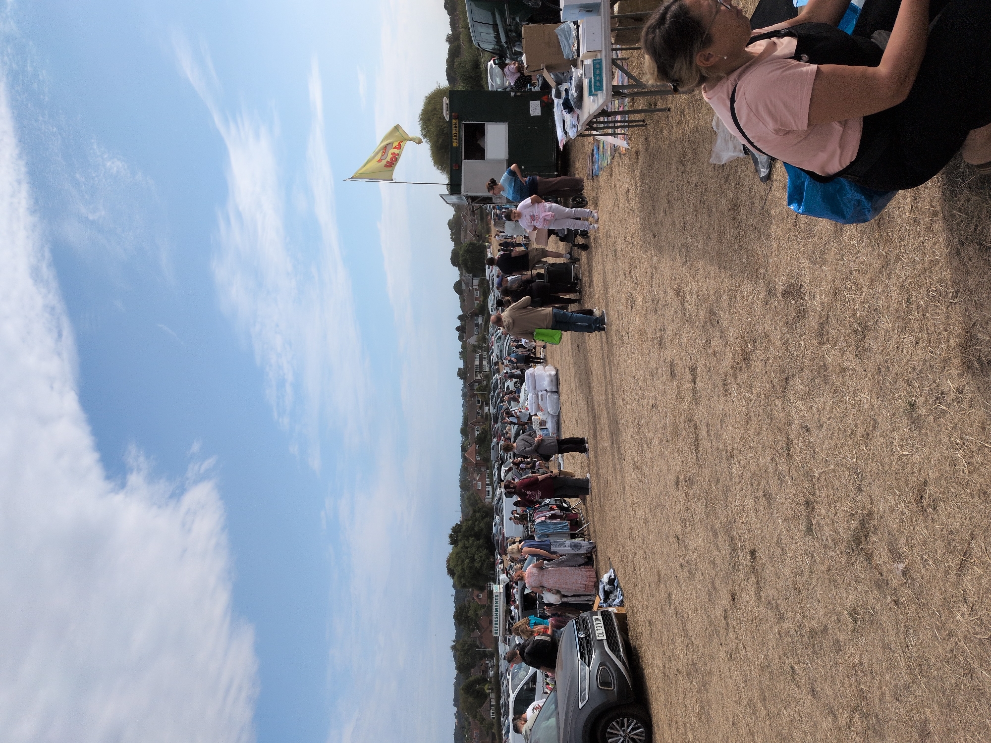 The image depicts an outdoor car boot sale, likely situated in a rural or semi-rural area within South Staffordshire, United Kingdom, given the presence of parked cars, stalls, and a natural landscape with trees and houses in the background. The scene is busy with numerous people browsing and selling goods.
In the foreground, on the right side, a woman wearing a pink t-shirt and black overalls is crouched down next to a blue bag. She appears to be attending to items on the ground. A table with various items, including boxes and what seem to be merchandise, is visible to her right.
Further into the image, a green stall with a yellow flag displaying "Hot Dogs" is positioned. People are gathered around various stalls displaying clothing, household items, and other goods. Cars are parked in rows, serving as makeshift display areas for vendors. The ground is covered in dry grass. The sky is blue with scattered clouds. The overall atmosphere suggests a typical weekend community event.