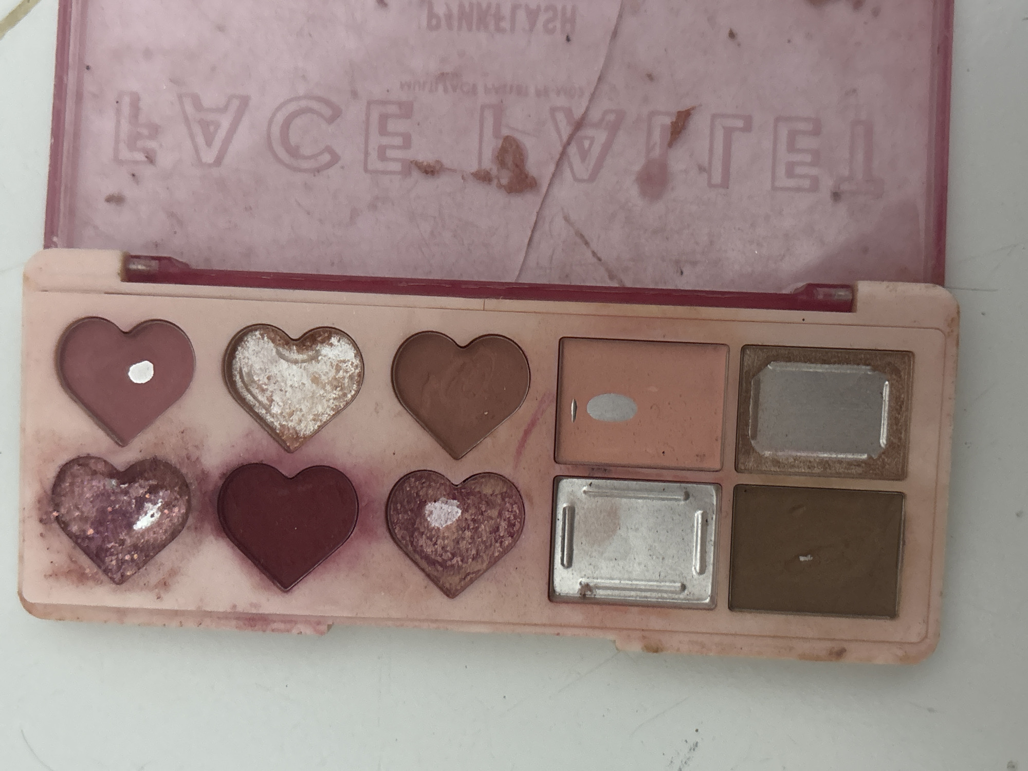 A pink eyeshadow palette with heart-shaped pans is shown in a top-down view. The palette is open, revealing the eyeshadow shades. The lid of the palette, which is also pink, is visible on the right side of the image and has the embossed text "FACETAL" and "PINKLYSH". The palette appears to be used, with some eyeshadow missing from the pans and dust and smudges on the surface. The background is a white surface, likely a table or countertop. The image was taken in North Jakarta, Indonesia.