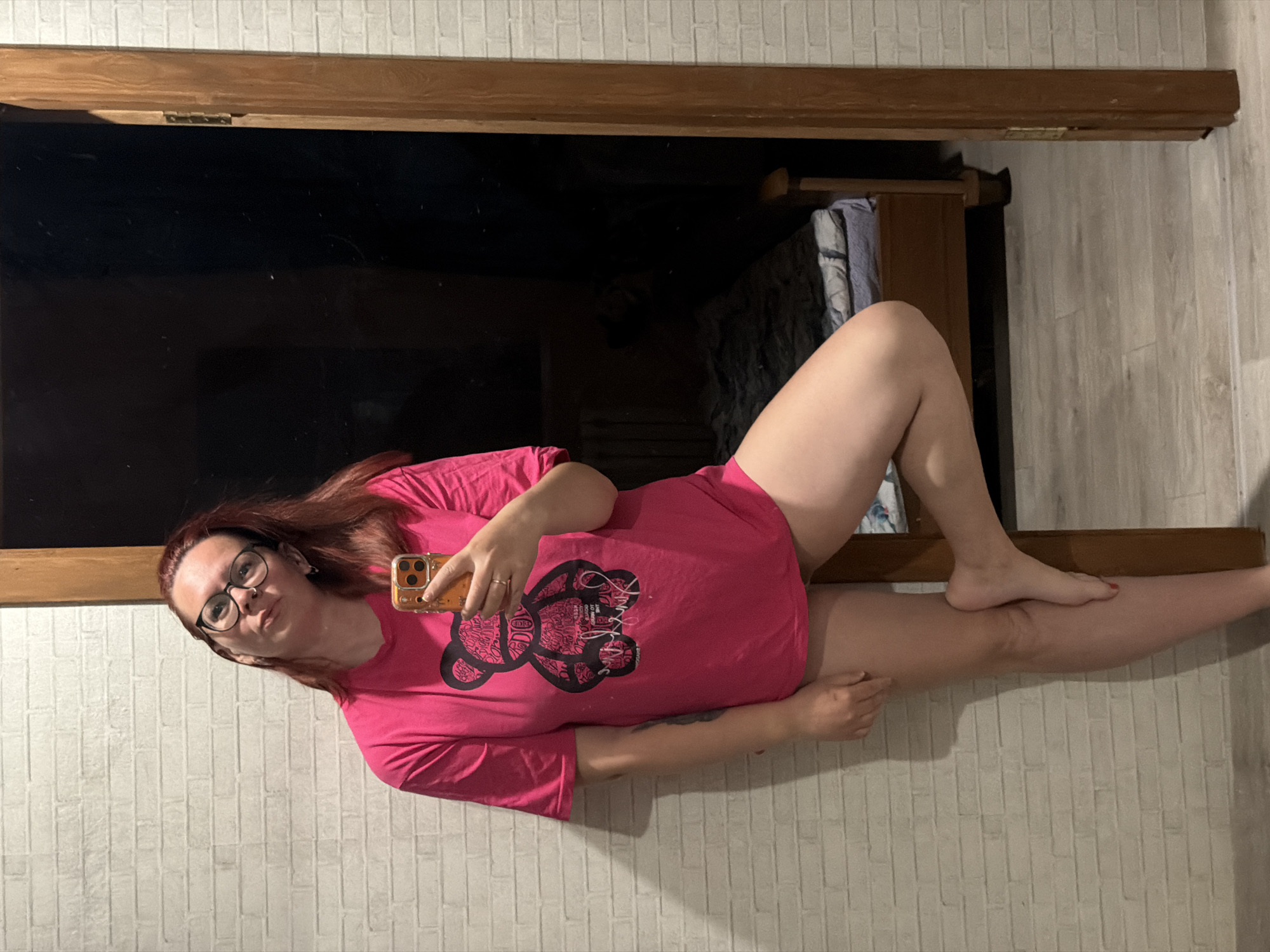 An adult female with red/auburn hair and black-rimmed glasses is depicted in a full-body mirror selfie, lying horizontally on what appears to be a wide window ledge or similar surface. She is wearing a bright pink t-shirt featuring a stylized bear graphic composed of intricate text, and pink underwear or shorts. Her bare legs are extended, one bent at the knee, with a foot resting on the other leg, showcasing red nail polish on her toes. Her left arm is bent, holding a smartphone with a yellowish case and multiple camera lenses, which she uses to capture her reflection in the dark window pane. Her expression is neutral, with a slight pout, as she looks towards the phone's camera.
The background consists of a light-colored brick-patterned wallpaper and a light wooden floor. A dark wooden frame borders the window, which reflects the dimly lit interior, showing hints of furniture or textiles inside. The text on the t-shirt's bear graphic includes highly stylized, small words, some resembling "D&G" and phrases like "CO GOOD" or "LOKING." The setting appears to be an indoor room, likely a home in Chaikovsky, Russia, possibly at night given the dark window reflection.