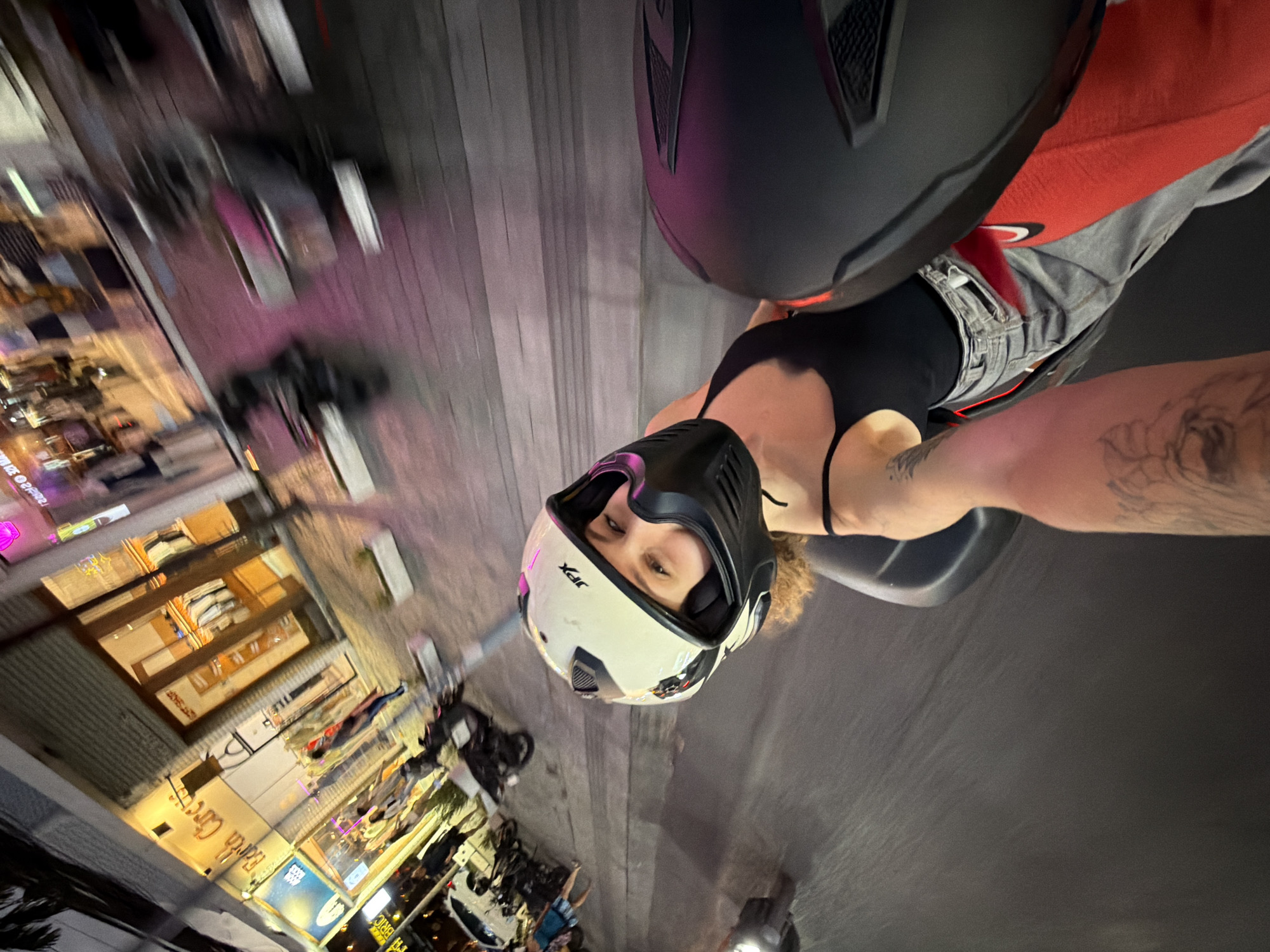 This is a selfie of two people on a scooter at night in North Kuta, Indonesia. The scooter is moving, causing motion blur in the background. The person in the foreground, facing the camera, is wearing a white helmet with black accents and a black face mask. They have curly hair and are looking at the camera. They are wearing a black tank top and gray jeans. The person taking the photo is holding the camera with their left arm, and their tattooed arm is visible. Another person's helmet, black in color, is prominently displayed in the lower right corner of the image. The background shows a street scene with blurred buildings, shops, and other scooters. The lighting is dim, with neon lights from the shops casting a pinkish hue on the street.