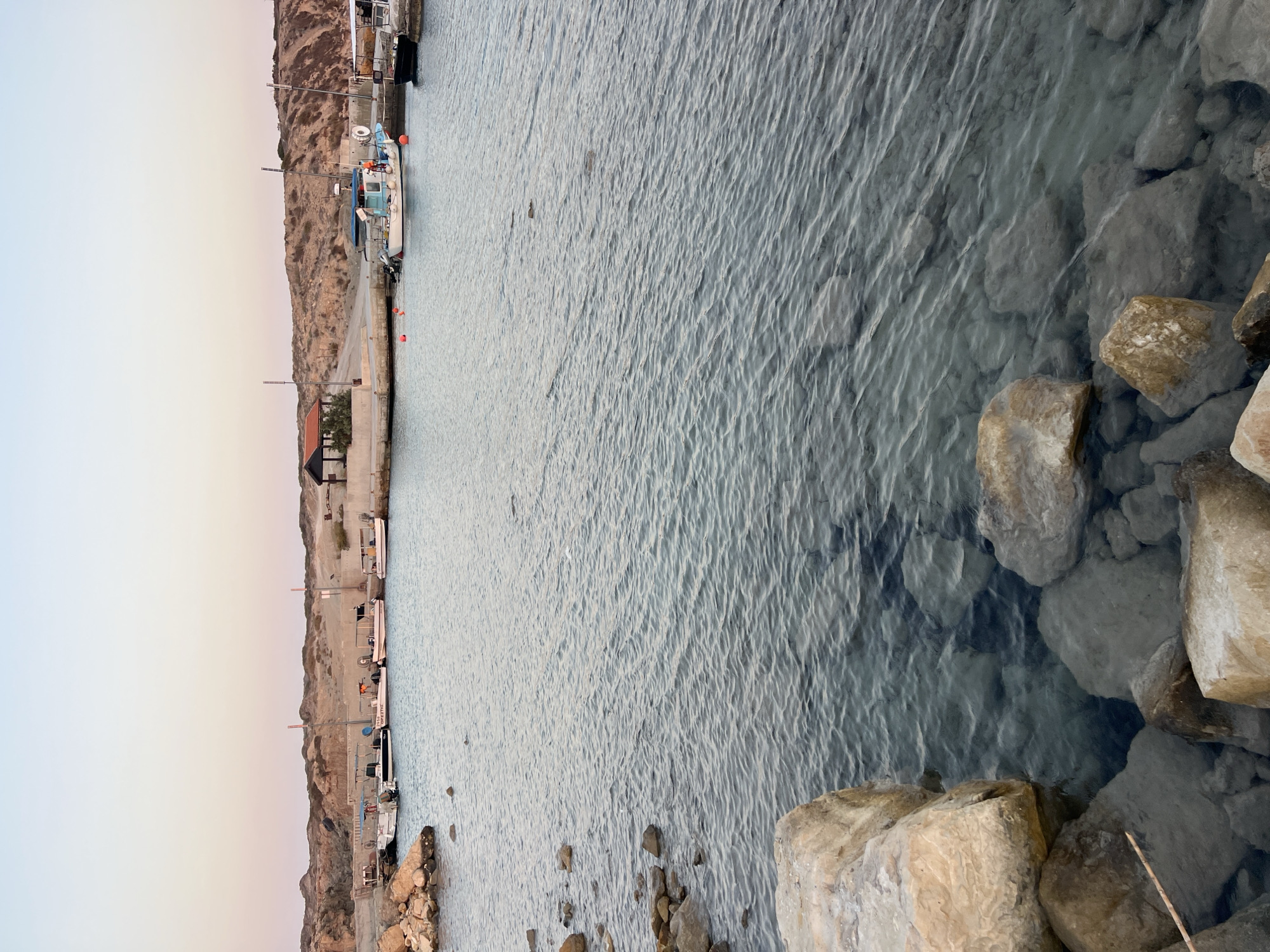 The image depicts a coastal scene at a harbor, likely in Akrotiri, as suggested by the location context. Several boats are docked along a concrete pier, with some appearing to be fishing vessels. The water is clear and calm, revealing rocks submerged beneath the surface near the shore. In the background, a rocky, arid hillside rises, characteristic of a Mediterranean landscape. The sky exhibits a gradient of pale blue and soft orange, indicating either dawn or dusk. The overall atmosphere is serene and quiet, with no people actively engaged in any visible activity. The time of day appears to be either early morning or late evening, due to the soft lighting and pastel colors in the sky. The clarity of the water and the rocky terrain provide specific geographical cues. One of the boats has visible text that reads "STAVROS".