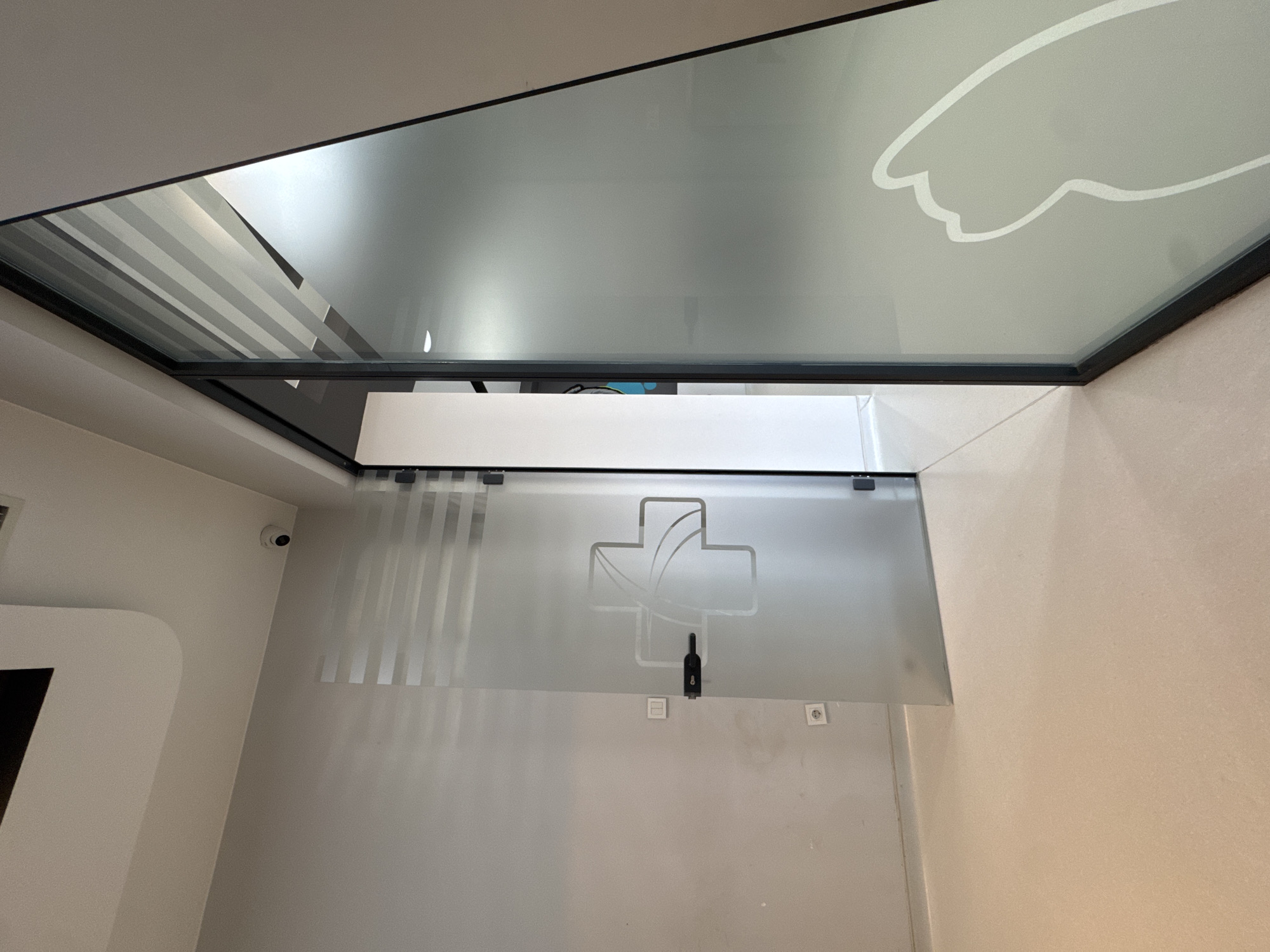 The image depicts an interior view, seemingly within a business or commercial establishment in Podgorica, Montenegro. The view is focused upwards, capturing a glass ceiling or skylight with a decorative design reminiscent of a cloud or abstract shape. Below this, there is a partial view of a frosted glass partition or door, adorned with a cross-like emblem containing leaf-like forms, commonly associated with medical or pharmaceutical services. The door has a black handle and a rectangular sensor below the emblem.
The surrounding walls are a light neutral color, and a surveillance camera is discreetly mounted on the wall to the left. There is a white structure, possibly part of a counter or display, partially visible on the left edge of the frame. Additionally, electrical outlets are visible on the wall below the glass partition. The overall impression is of a modern and functional space, likely a pharmacy or clinic, with attention to design elements.