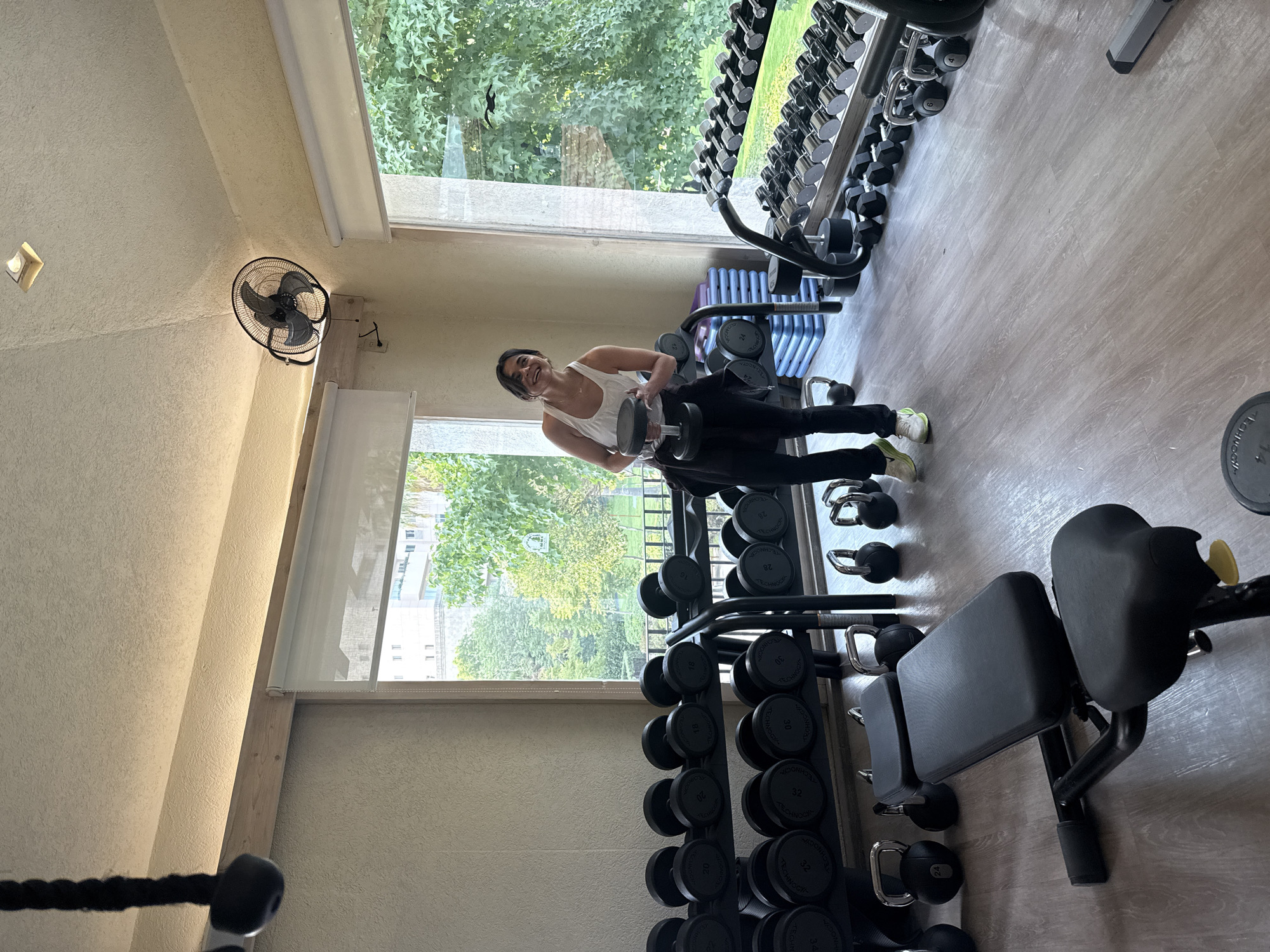 The image depicts a woman in a gym, engaged in a weightlifting exercise. She is positioned between a rack of dumbbells and a large window that looks out onto a view of greenery and buildings, possibly an urban park or residential area. The gym appears well-equipped with various types of weights, including dumbbells and kettlebells, and a weight bench. A wall-mounted fan is visible in the upper left corner. The background suggests the location could be the city of Huixquilucan de Degollado, Mexico, given the architecture and landscape visible through the window. The woman is wearing a white tank top and black leggings. She is holding dumbbells in both hands, executing a specific weightlifting move.