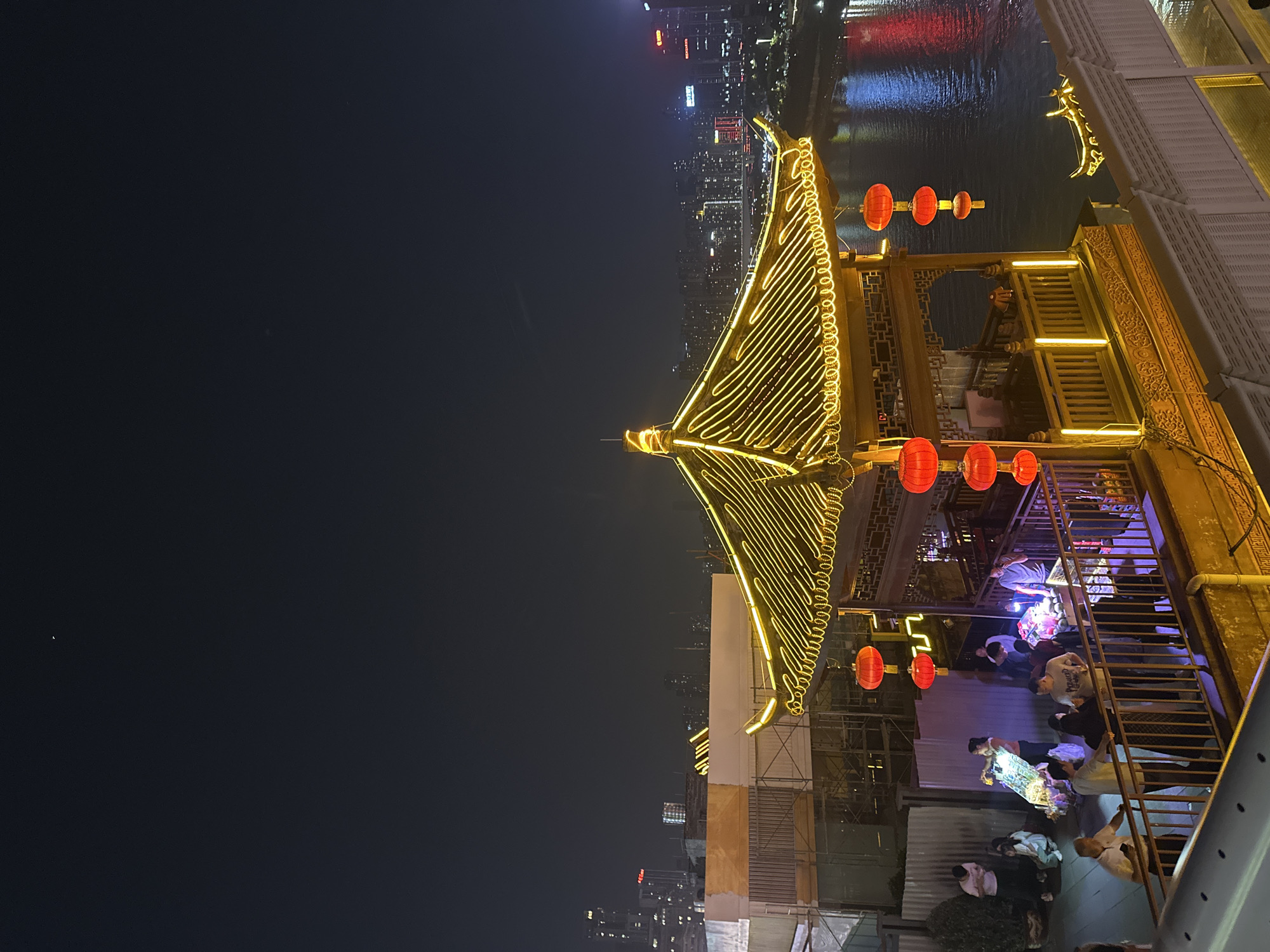 The image captures a vibrant night scene in Yuzhong District, China, showcasing a blend of traditional architecture and a modern cityscape. A prominent, ornate structure, resembling a traditional Chinese pavilion or a boat with such a superstructure, dominates the foreground. Its distinctive curved roof is intricately outlined with bright yellow LED strip lights, creating a warm, glowing effect against the dark night sky. Multiple red lanterns are suspended from the eaves of this structure and other nearby elements, adding to the traditional aesthetic.
To the right, a dark body of water, likely a river, reflects the distant city lights in vibrant streaks of red, blue, and white. Across the river, the background reveals a dense modern city skyline, composed of numerous illuminated high-rise buildings.
In the lower right portion of the image, below the main illuminated structure, several people are gathered on what appears to be a multi-level outdoor or semi-outdoor area, possibly a restaurant or social venue. They are seen standing and interacting in groups, suggesting a lively social event or evening leisure activity. A metal staircase with railings is visible within this area. The overall atmosphere is festive and bustling, illuminated by a combination of decorative lights and the ambient glow of the city. No discernible text is clearly visible in the image.