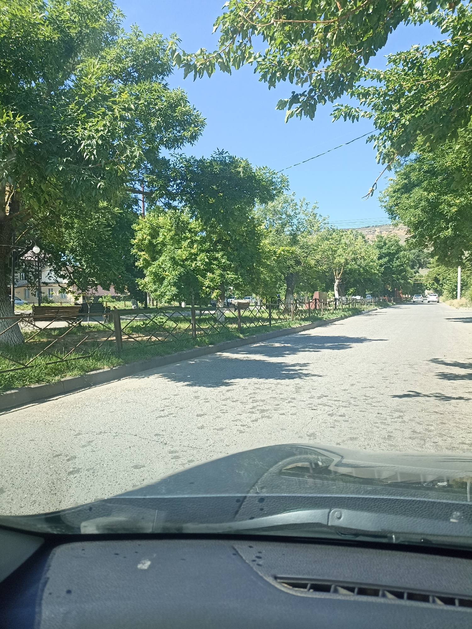 The image shows a street in Izberbash, Russia, on a sunny day. The street is lined with trees and a fence. There are a few cars on the street and a building in the background. The perspective is from inside a car, looking out. The dashboard and windshield of the car are visible in the foreground.