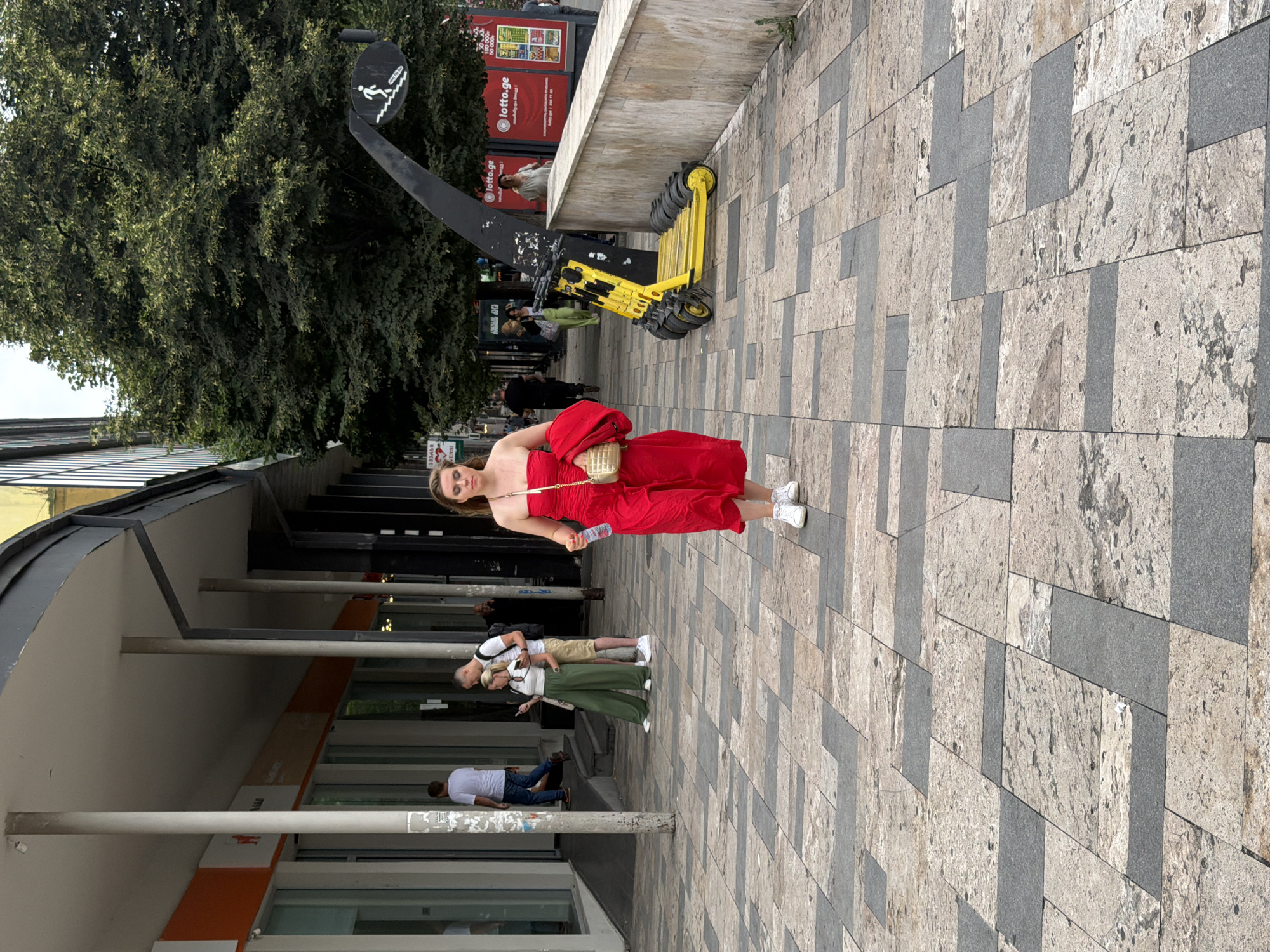 A woman in a red strapless dress walks towards the camera on a patterned pedestrian walkway in Tbilisi, Georgia. She carries a water bottle in her right hand and a small gold purse on her left shoulder. She is wearing white sneakers and a red jacket draped over her left arm. Behind her, a couple stands near a storefront, the man looking at his phone while the woman, in green wide-leg pants and a white top, looks at her phone. Further back, other pedestrians are visible walking along the street, and a row of yellow electric scooters is parked near a building with advertisements for Lotto.ge. Trees and a tall building form the background of the urban scene.