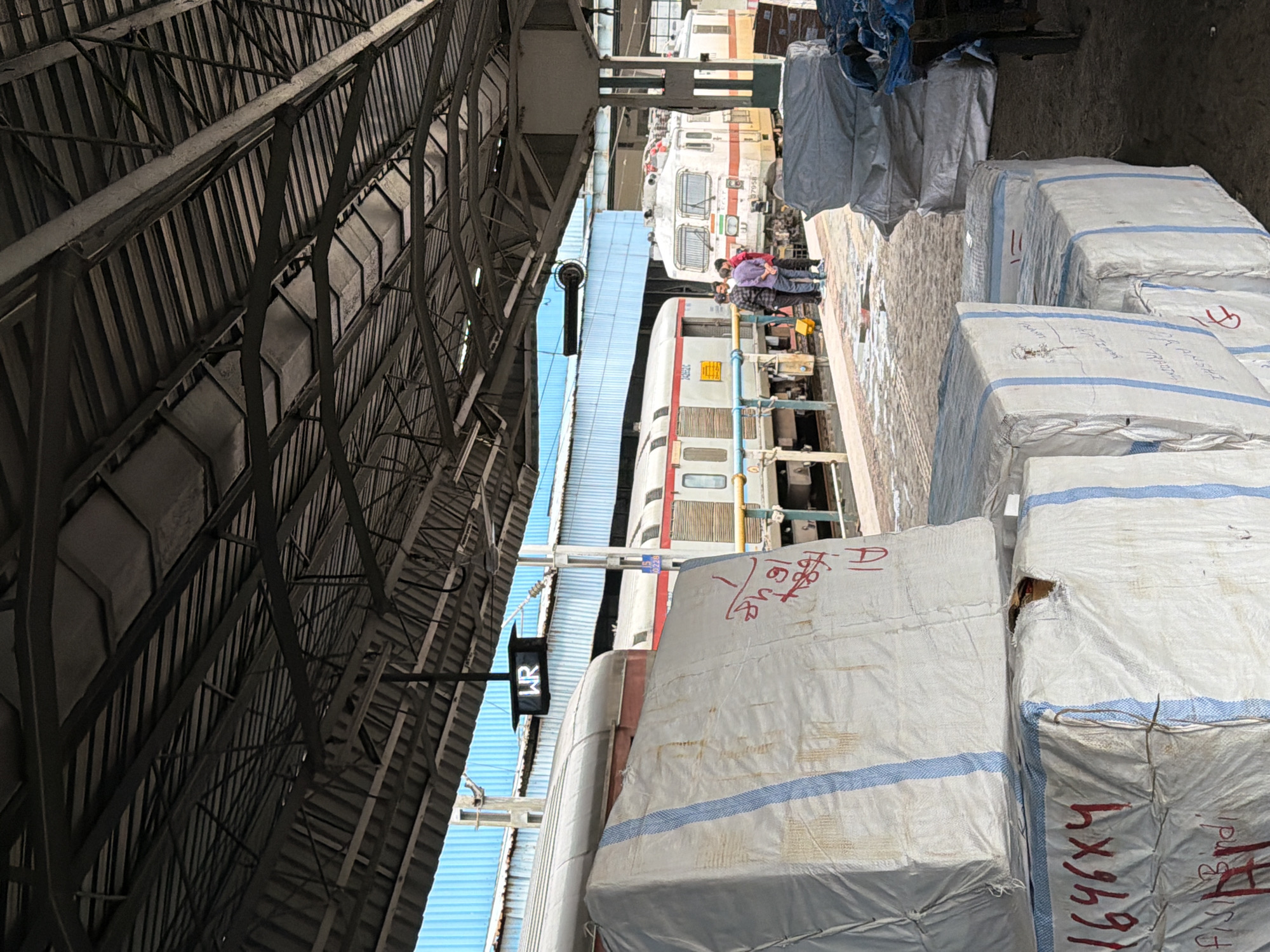 A train station platform in Mumbai, India is depicted, covered by a large metal roof supported by beams. In the foreground, several large rectangular packages wrapped in white material with blue stripes are stacked. Red markings are visible on some of the packages. In the middle ground, a train is stopped at the platform. Three men are standing near the train, appearing to be in conversation. A sign with the letters "WR" is hanging from the roof. In the background, more train cars are visible, along with the blue siding of a building. The platform area appears to be a cargo or logistics hub within the train station.