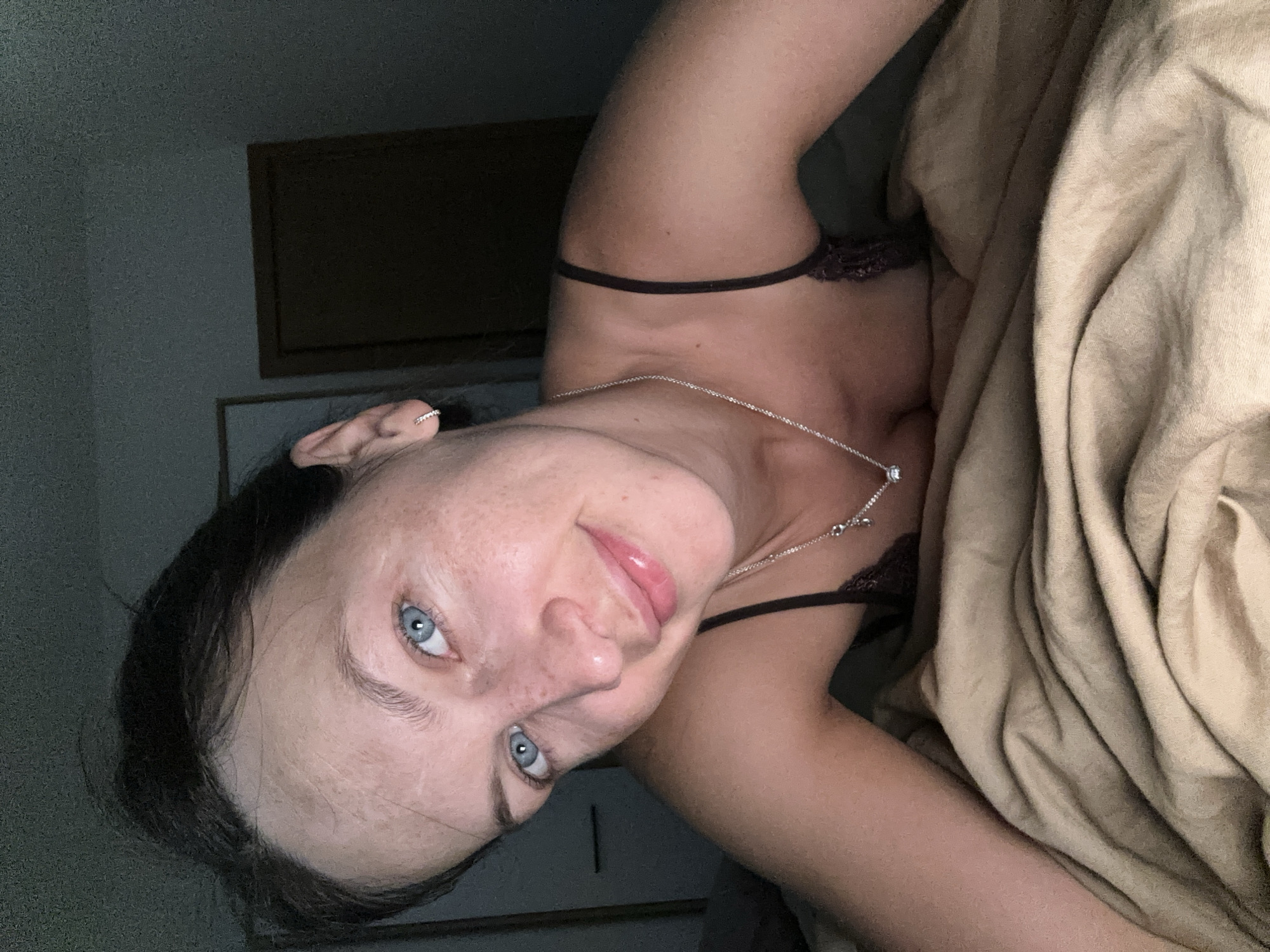 A woman with blue eyes and freckles lies on a beige bed. She is wearing a dark camisole and a silver necklace. Her hair is dark brown and pulled back. The background is dark, with a hint of a doorway and a wall. The image is an indoor selfie taken at night.