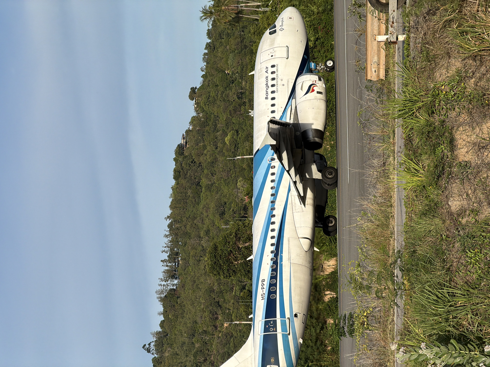 An exterior, eye-level, medium shot captures a Bangkok Air airplane taxiing on a runway in Baan Bo Phut, Thailand. The airplane, predominantly white with blue accents and stripes, features the text "Bangkok Air" and "ASIA'S BOUTIQUE AIRLINE" on its fuselage. The registration number "HS-PPB" is visible on the tail. The aircraft's engines are clearly visible, as are its landing gear. The runway is paved and marked with white lines.
In the background, a lush, green hillside is covered with dense trees. A few houses or structures are sparsely visible nestled within the foliage on the hill. The sky is clear and a light blue, with some faint cloud streaks. In the foreground, the edge of the runway is lined with dry grass, weeds, and some foliage, suggesting a natural, undeveloped area surrounding the airport. A rusty guardrail is present along the side of the runway in the lower right corner of the frame. The lighting indicates it is daytime, with the sun casting shadows and highlighting the aircraft. No people are visible in the image.