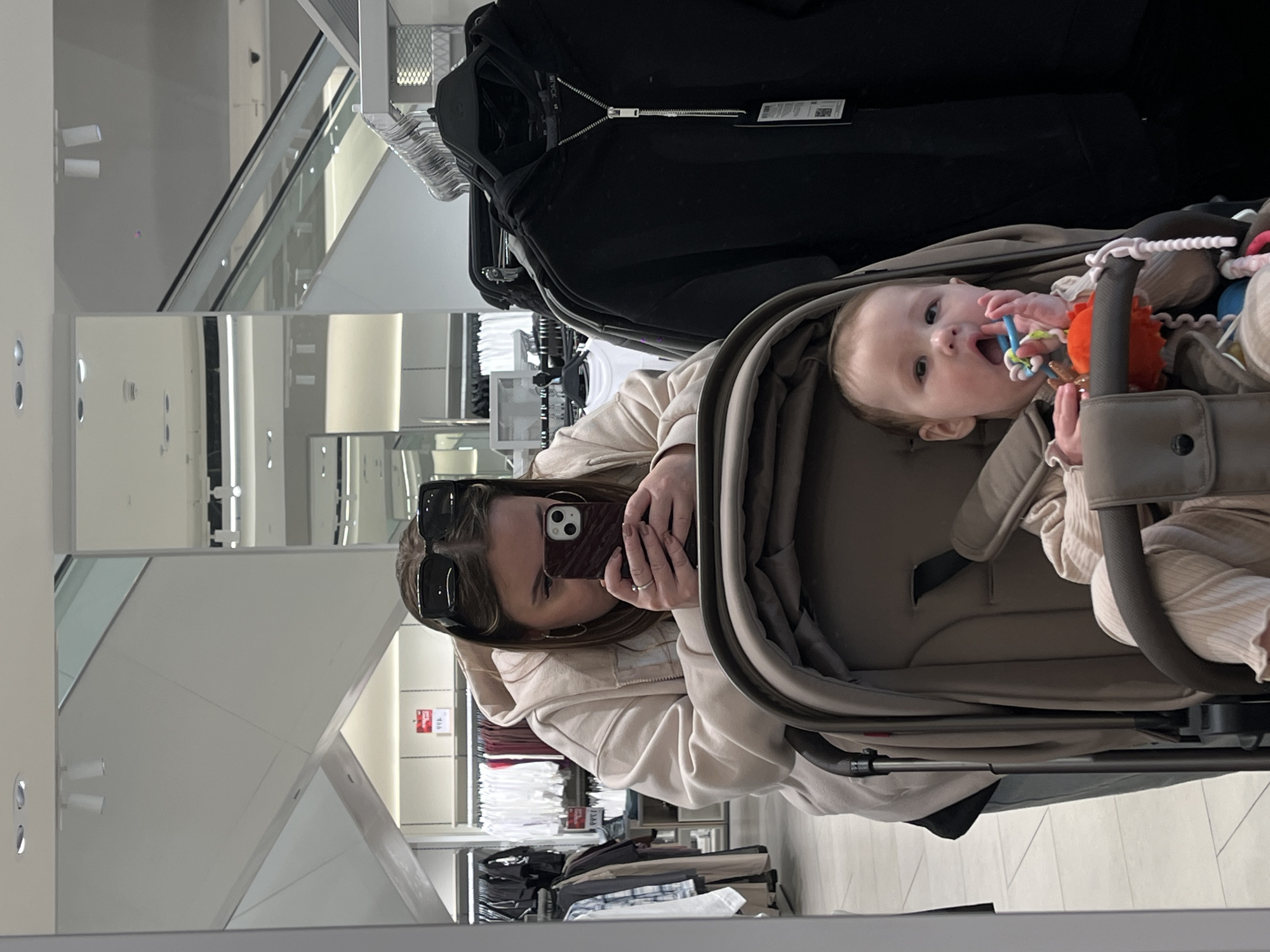 This mirror selfie, taken in Krasnodar, Russia, captures a woman and a baby in a brightly lit retail environment. The image is horizontally oriented, showcasing the reflection in a large mirror.
In the center, a woman with long, light brown hair and sunglasses perched on her head holds a smartphone to take the photo, partially obscuring her face. She is wearing a light beige hooded top and a ring on her left ring finger. To her right, a baby is seated in a taupe-colored stroller, looking towards the camera. The baby has light hair and is actively chewing on a colorful teether toy, which includes blue, orange, and white elements.
The background, reflected in the mirror, reveals a modern clothing store. Various clothing items are displayed on racks, including a prominent black zippered jacket with a visible tag that reads "BLACK". In the deeper background, rows of apparel on hangers are visible, along with clean white walls and subtle light fixtures. An escalator or set of stairs with glass railings is reflected, indicating a multi-level store or shopping mall. A red sign, partially obscured, shows numbers like "499" or "4999," suggesting pricing or sale information. The overall scene depicts a casual shopping outing during the daytime.