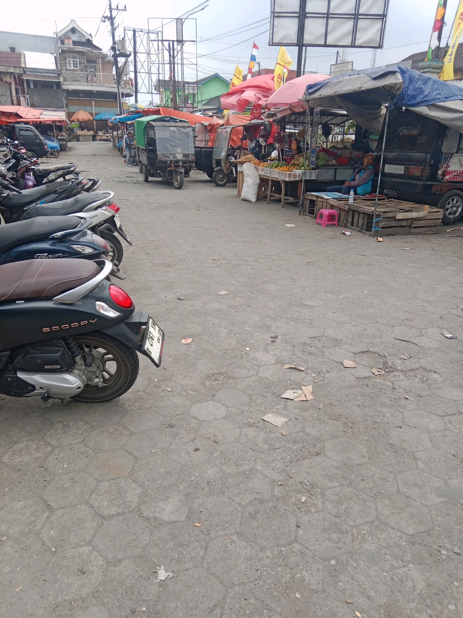 The image depicts a street-level view of a market in Batur, Indonesia. In the foreground, several parked motorcycles are visible, with the closest being a black Honda Scoopy. The street surface is paved with hexagonal interlocking concrete blocks, showing signs of wear and scattered debris. To the right, a market stall is set up, covered by a blue tarp and selling various fruits, including oranges and what appear to be watermelons. A person, likely a vendor, is seated on wooden pallets under the stall, wearing a blue jacket and a cap. Behind the stall, a black truck is parked. Further down the street, more market stalls are visible, some with red and orange awnings, and a few three-wheeled vehicles, possibly for transport or market goods. Buildings, some with multiple stories, line the left side of the street, with architectural elements suggesting a tropical or Southeast Asian context. Utility poles and wires are present in the background. The overall impression is a busy, open-air market environment with a mix of vehicles, vendors, and goods.