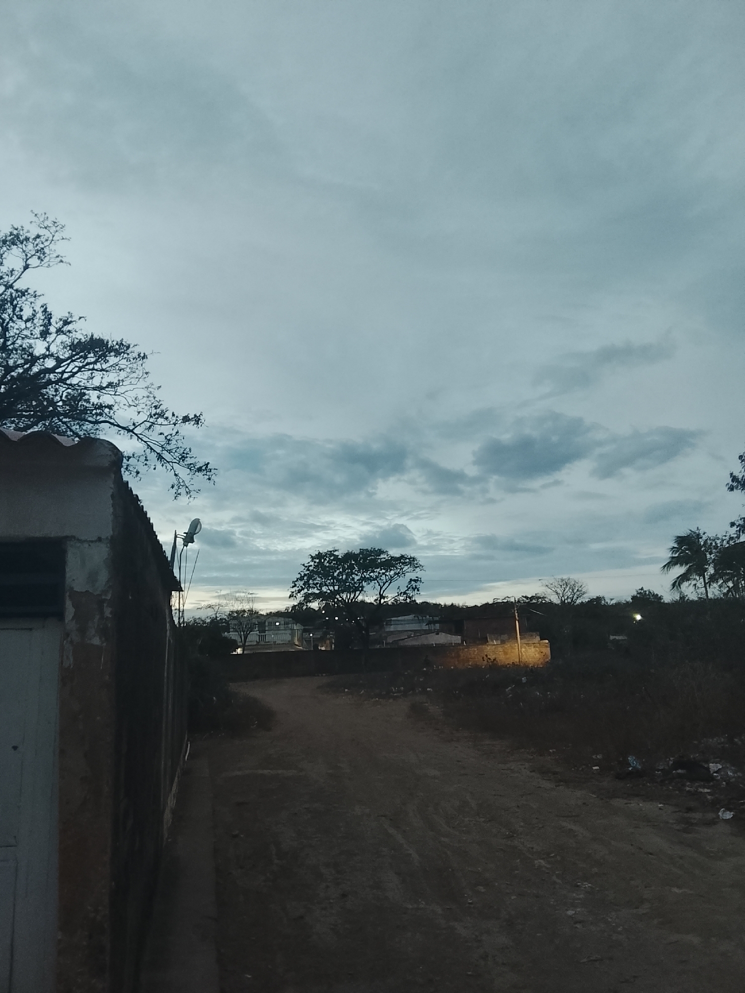 The image depicts a dirt road in what appears to be a residential area. On the left is a rundown building with a partially visible door and a corrugated metal roof. A satellite dish is mounted on the building. A dirt road stretches into the distance, flanked by vegetation and buildings. Light suggests it's either dusk or dawn. The sky is overcast with heavy clouds. The specific location in Juan Griego, Venezuela is not definitively discernable from the image alone. There is no indication of people present in the image.