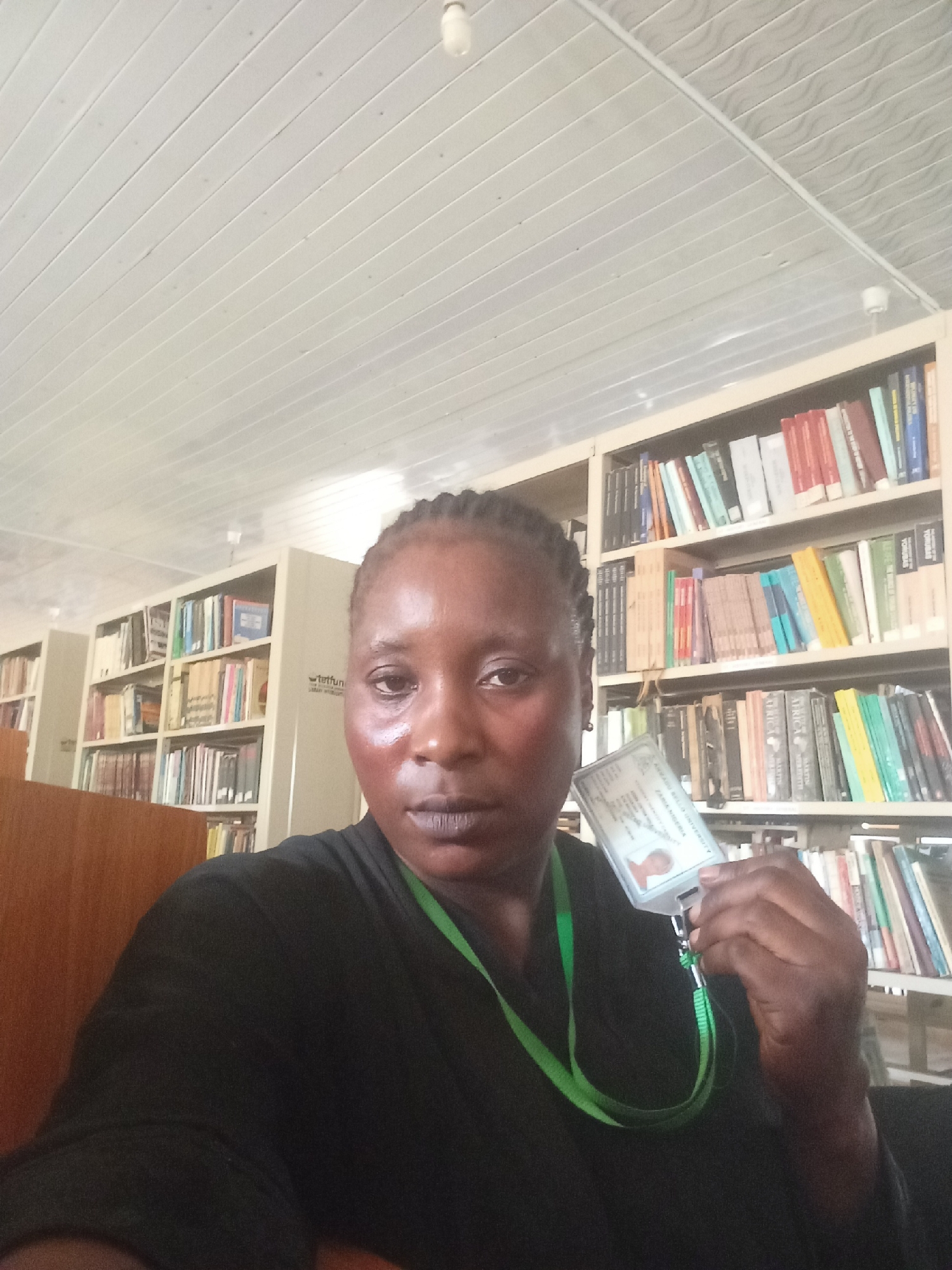 A woman is taking a selfie indoors, presumably in a library in Akwanga, Nigeria. She is holding up an identification card. The background shows multiple bookshelves filled with books. The ceiling has a white, paneled design. The woman is wearing a black shirt and a green lanyard.