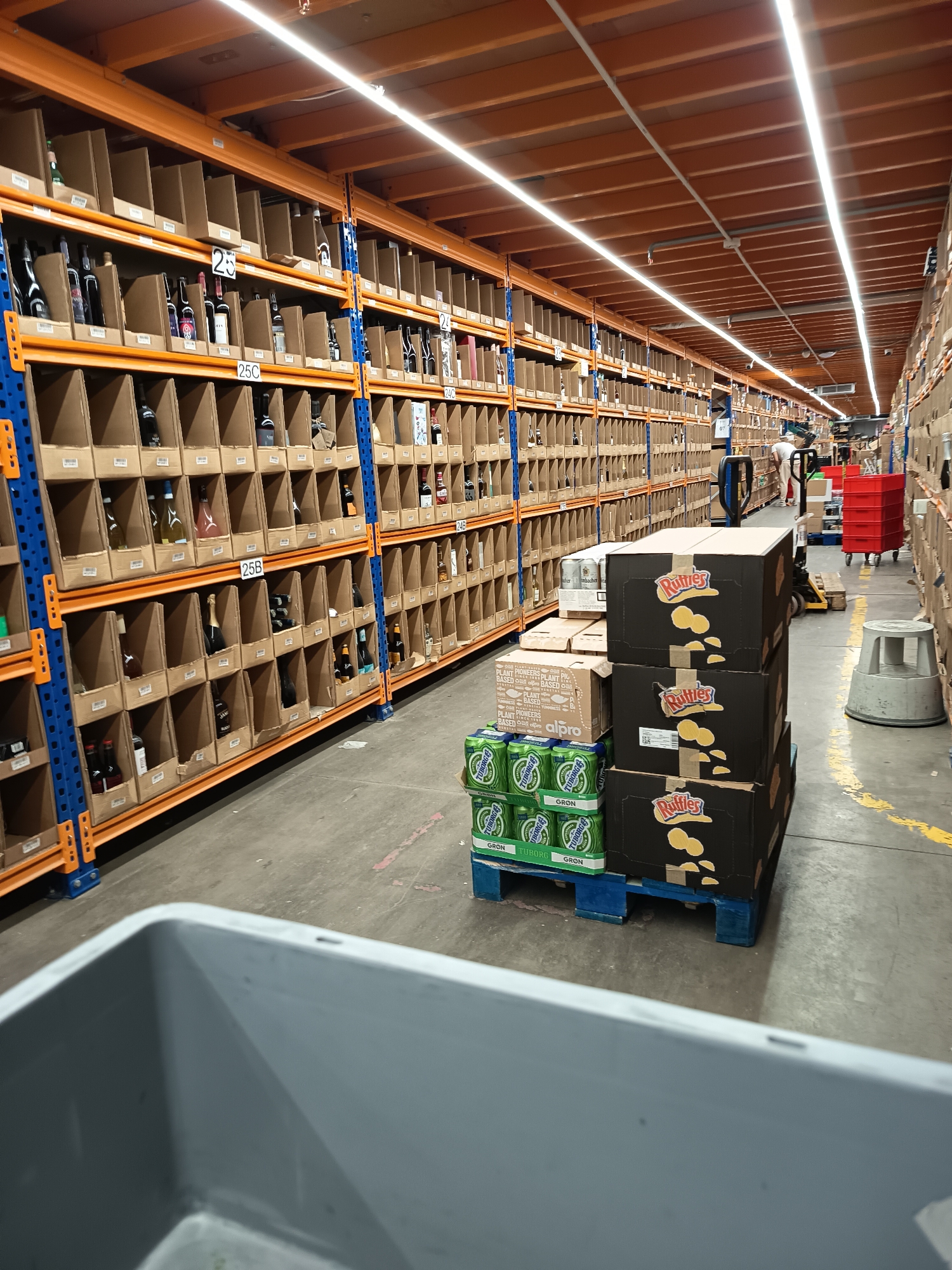 The image shows the interior of a warehouse in Sofia, Bulgaria. Rows of orange shelving units, filled with individually boxed items, line the left side of the frame. The shelves are labeled with numbers such as "25", "25C," and "25B." A pallet loaded with boxes of "Ruffles" potato chips, cartons of "alpro" plant-based drinks, and packs of "Tuborg Gron" beer is centrally positioned. In the background, a worker in a white shirt operates a piece of machinery, and a red cart is visible. A gray plastic bin is in the foreground. The warehouse is illuminated by long, ceiling-mounted lights.