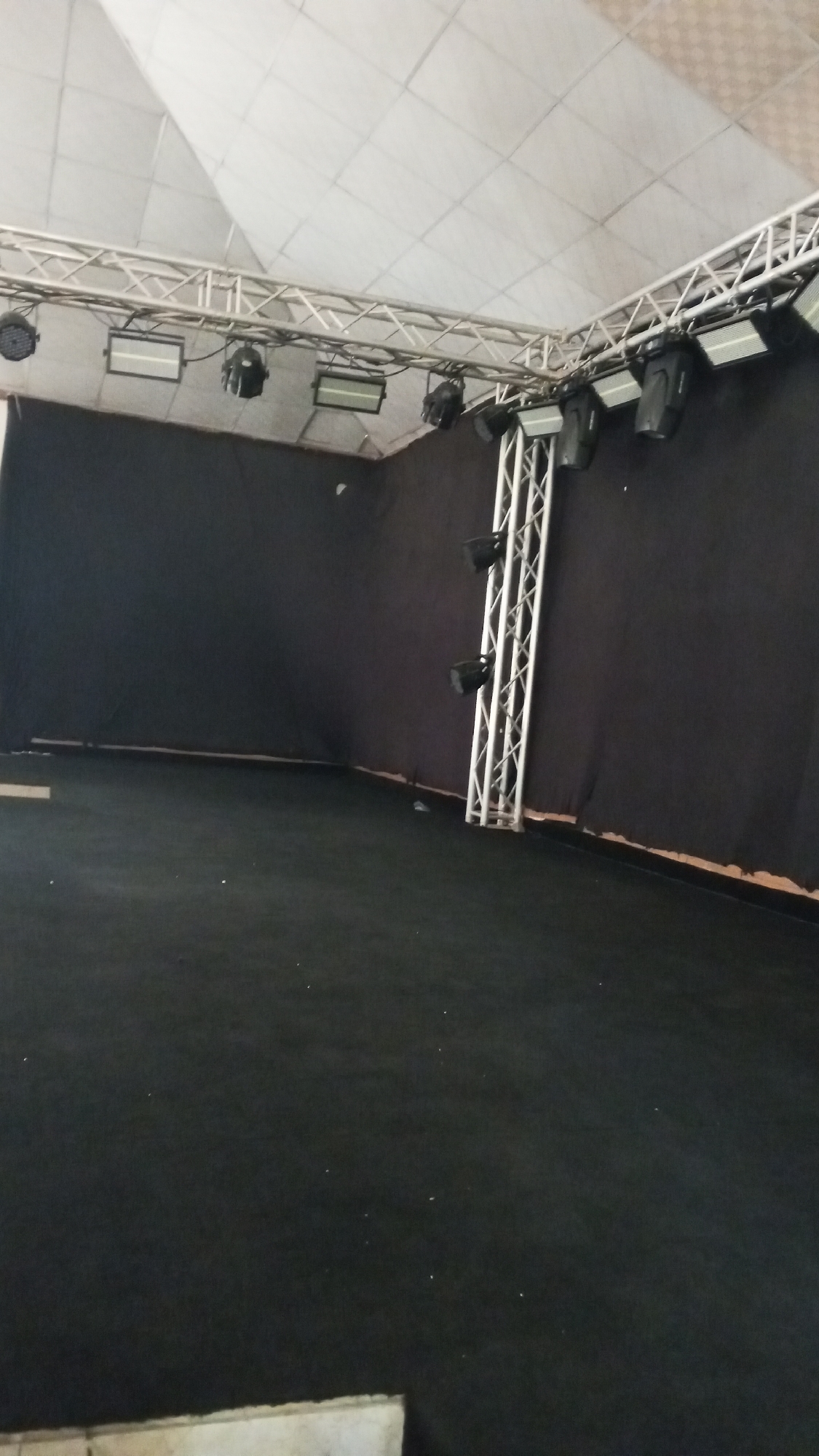 The image shows an empty indoor stage setup. The stage floor is covered in black carpet. Behind the stage, a black curtain backdrop is hung. Above the stage, a white fabric ceiling is visible, with a metal truss system supporting several stage lights. The truss is silver and triangular in shape. The lights appear to be professional stage lighting fixtures, some are rectangular with white fronts, and others are round and black. A silver truss structure is also present on the right side of the stage, rising vertically and supporting more stage lights. The overall impression is of a prepared space for a performance or event, but it is currently unoccupied. There are no people visible in the image. The lighting suggests it could be daytime or an artificially lit indoor space. No weather or specific location cues are evident, making the location unknown. No text is visible in the image.