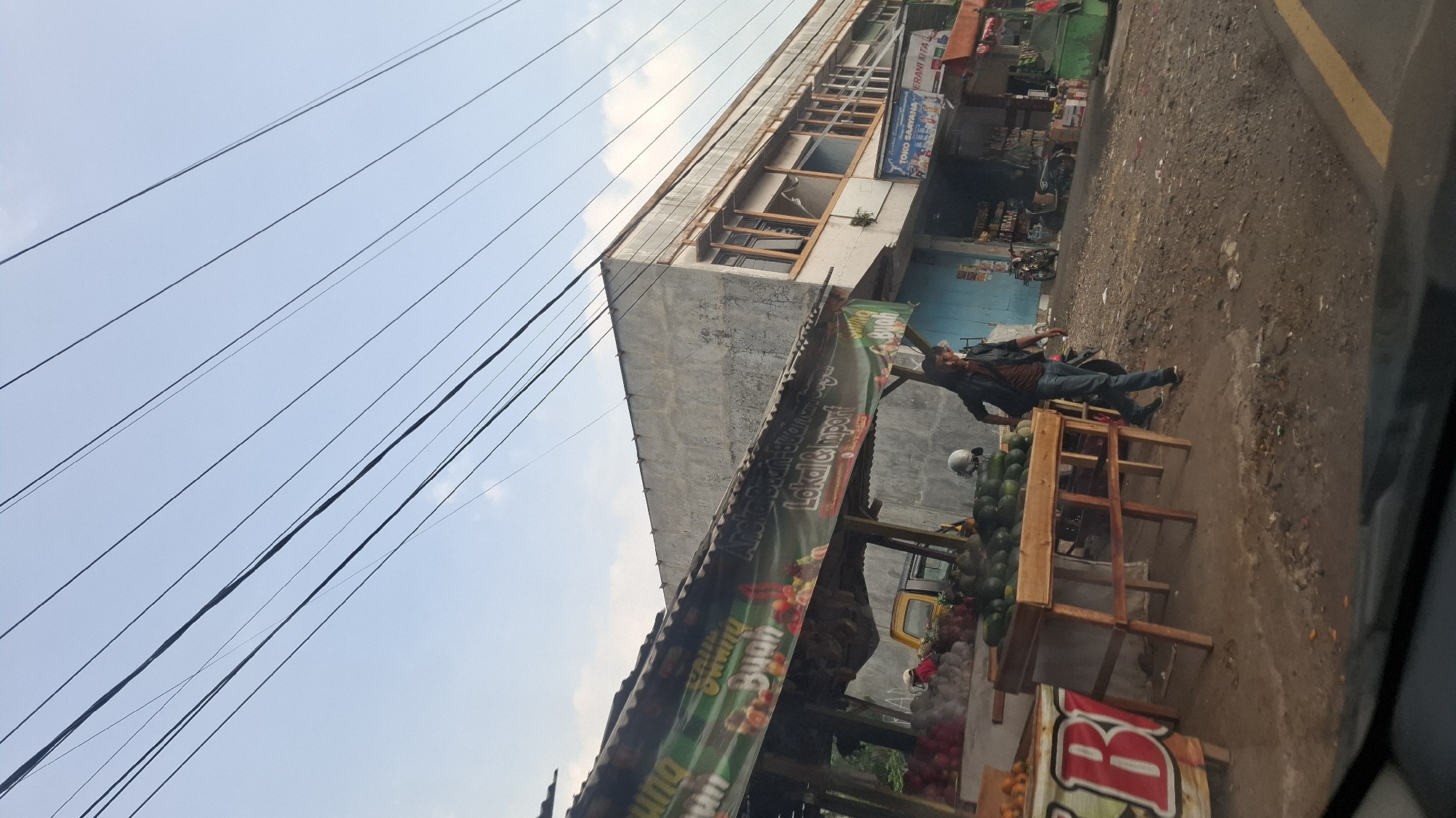 In the city of Cilaku, Indonesia, a man in a dark jacket and blue jeans walks past a fruit stand. The stand has a tarp overhead and displays a variety of fruits, including watermelons and what appear to be avocados. Behind the fruit stand is a building with a concrete facade and windows. Overhead, power lines crisscross the sky. The ground is dirt and gravel, with some debris scattered around. A yellow truck is visible in the background. The overall scene depicts a street-side market.