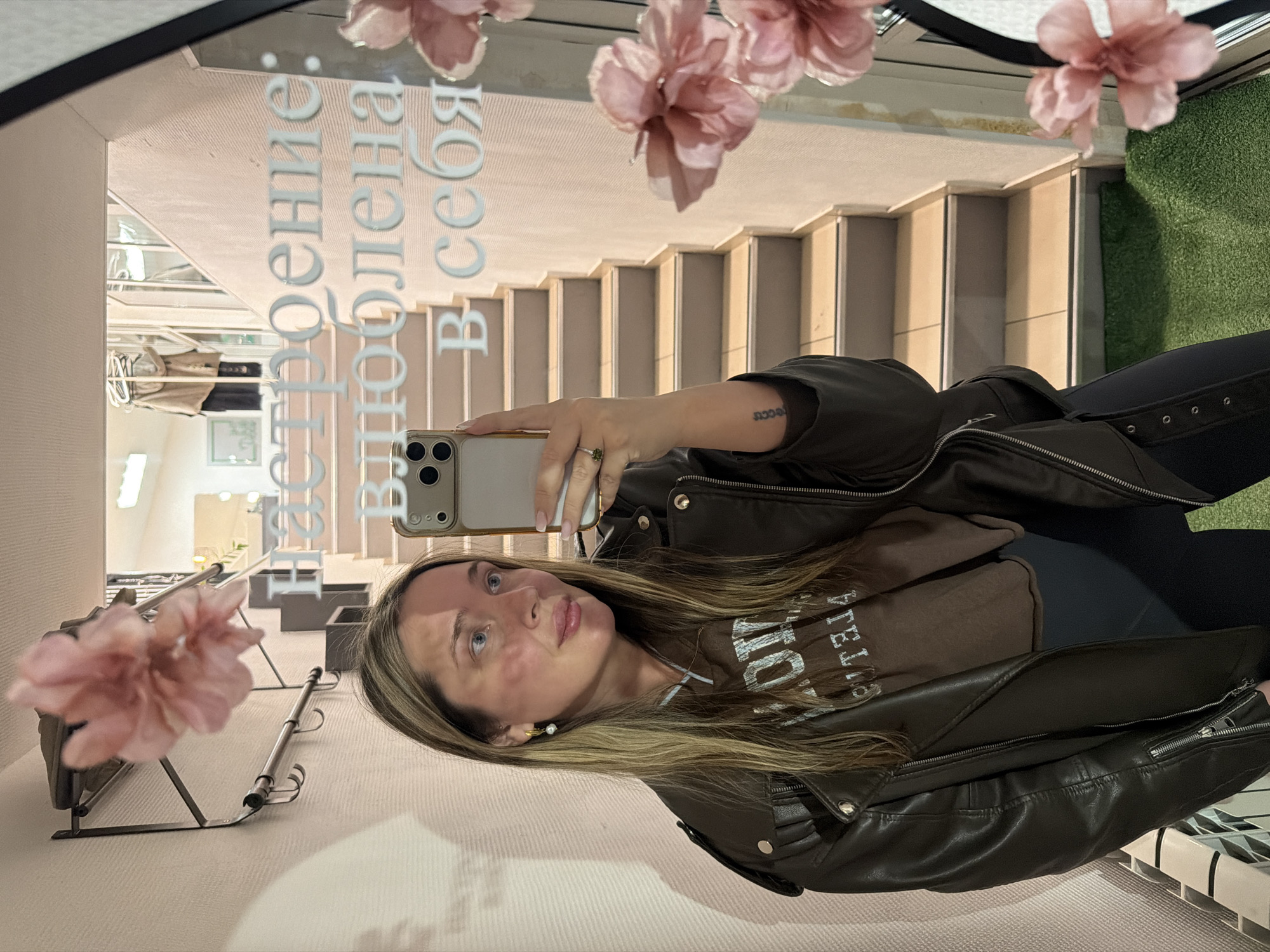 A woman is captured in a mirror selfie, looking slightly upwards and to her left with a neutral expression. She has long, straight blonde-brown hair and blue eyes. She wears a dark brown leather jacket over a brown hooded sweatshirt with white text (partially visible as "STELLA" or similar lettering) and dark pants. Her right hand holds a silver iPhone, taking the photo. A ring with a green stone is visible on her left ring finger, and a small tattoo, reading "נשמה" (Neshama, Hebrew for 'soul'), is visible on her left forearm. A gold hoop earring with a pearl hangs from her right ear.
The background reflected in the mirror shows an indoor setting, possibly in Chaikovsky, Russia. A light-colored staircase descends in the upper-right portion of the reflection, while a patch of artificial green turf is visible at the bottom right. To the far left, a metallic clothing rack holds some light-colored garments. Decorative artificial pink flowers are taped to the top edge of the mirror, with two on the left and one on the right.
White Cyrillic text is written vertically on the mirror, appearing horizontally across the top-left when viewed correctly: "Настроение: расположена в себя" which translates to "Mood: inclined in herself" or "Mood: self-disposed." The lighting is soft and even, suggesting an indoor setting without strong natural light sources.