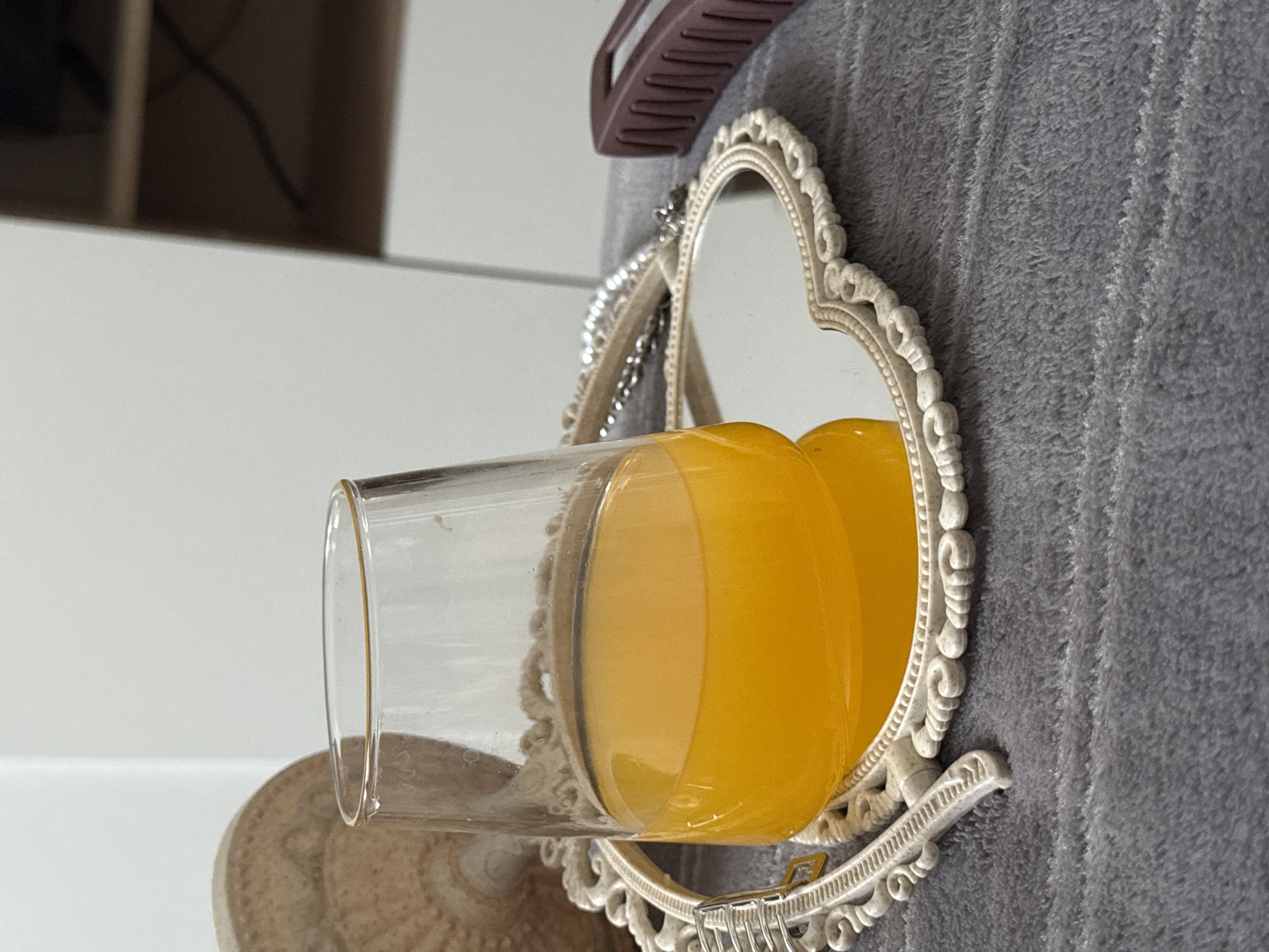 A glass of orange juice is reflecting in a small, white heart-shaped mirror. The mirror is resting on a surface and has a grey, textured background. A hair clip is visible above the mirror. The location appears to be indoors in the city of Unknown, Unknown.