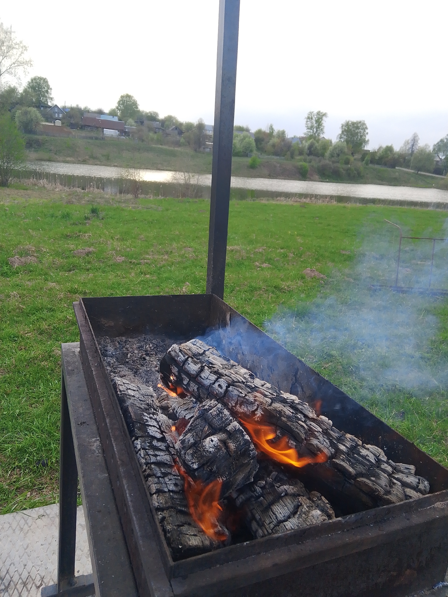 The image shows a charcoal grill containing burning logs in an outdoor setting, potentially in or near the city of Фоки, Russia, as indicated by the query context.
The grill is a metallic rectangular structure with visible ash and partially burnt logs emitting flames and smoke.
The background includes a grassy area, a body of water, and a distant view of buildings and trees, indicating a rural or suburban setting. The environment appears to be relatively quiet and still. The lighting suggests it is daytime, with overcast conditions.