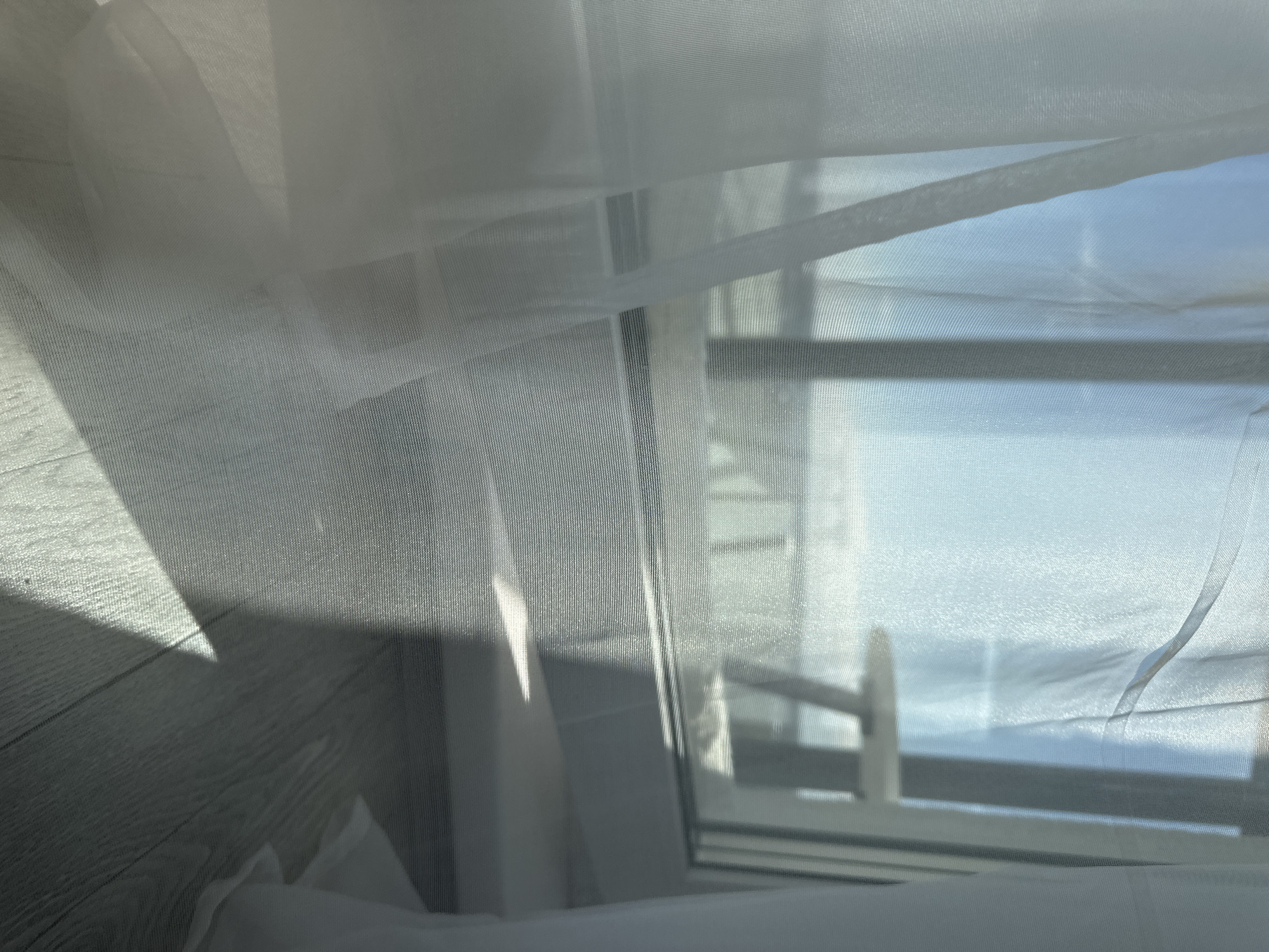 A view through a window with a sheer white curtain, in what appears to be a room with light-colored, wood-patterned flooring. Outside, a clear sky is visible along with a body of water. There's a window frame and what appears to be part of a railing or structure beyond. A combination of light and shadow is present on the curtain and the flooring. The city and location of Unknown, Russia are not visibly discernible.