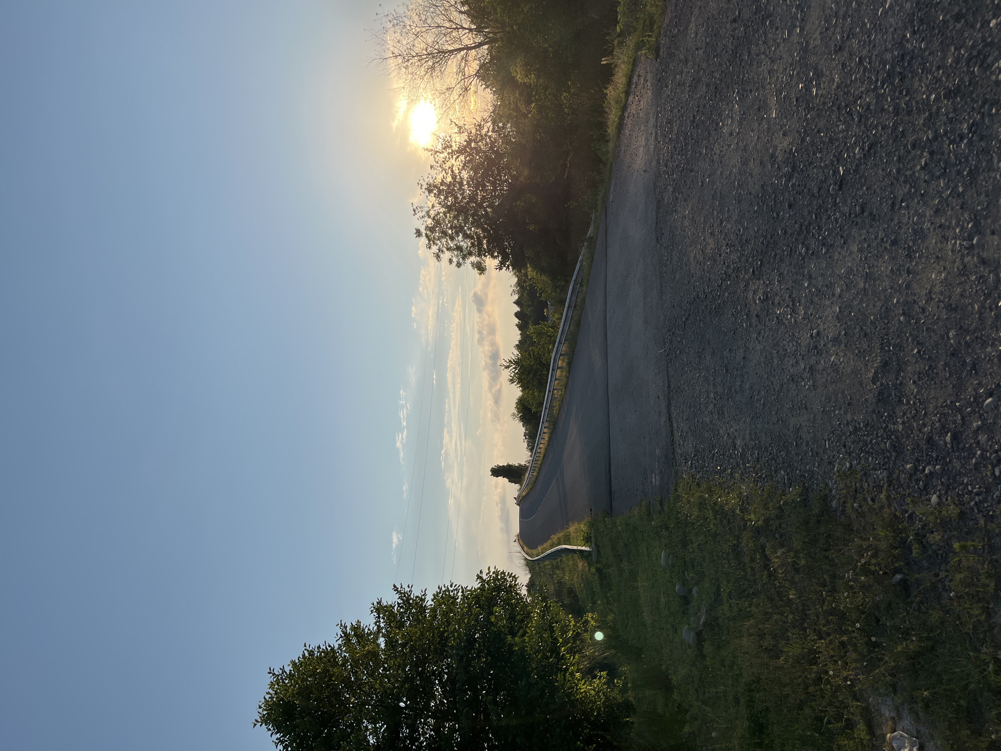 The image depicts a road ascending a gentle incline, bordered by a metal guardrail on the right and a lush, green embankment with scattered rocks and vegetation on the left. Tall trees are situated on either side of the road, with dense foliage. The sky is a clear blue, transitioning to a softer hue near the horizon where a bright sun is setting, casting a warm glow and partially obscured by clouds and trees. A light haze is visible in the distance. The road surface is asphalt with a gravel shoulder. The scene appears to be a rural or semi-rural setting, possibly on the outskirts of Ureki, Georgia, given the natural landscape and clear skies typical of the region during sunset. There are no people or vehicles visible in the frame.