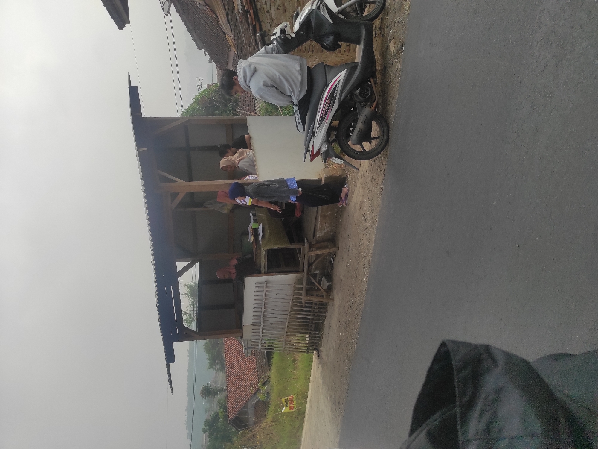 The image depicts a roadside scene in a rural area, likely in or around Sumedang, Indonesia, characterized by overcast skies and a somewhat hazy atmosphere. In the foreground, a dark asphalt road occupies the lower portion of the frame, with a patch of disturbed dirt and gravel to its right.
Dominating the mid-ground is a simple, open-sided roadside stall or kiosk constructed with a corrugated metal roof supported by wooden beams. The stall appears to be a vendor's spot, with a counter and shelves visible inside. A white wooden fence, appearing somewhat dilapidated, encloses a portion of the stall. A wooden bench is placed in front of the fence.
Several people are present at and around the stall. A woman wearing a pink headscarf is visible behind the counter, possibly serving or attending to customers. A young person wearing a blue headscarf and dark clothing stands near the counter. Another woman, also wearing a headscarf, is seen inside the stall. Two other individuals, partially obscured, are also visible within the stall area.
To the right of the stall, a white and black scooter is parked. A young man wearing a grey hooded sweatshirt is seated on the scooter, looking to his left. The scooter has some decorative graphics on its side.
The background reveals a hilly landscape with scattered houses featuring red tile roofs. Vegetation, including trees and grassy areas, is prevalent. A yellow sign with text is visible on a post near the road to the left of the stall. The overall impression is a quiet, functional scene in a developing or rural setting, likely a point of commerce for the local community.