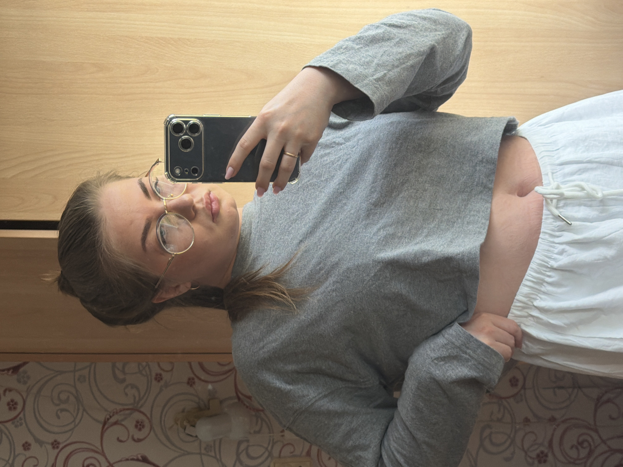 A young woman is taking a selfie in what appears to be a bathroom or bedroom. She is wearing round-rimmed glasses, a gray long-sleeved crop top, and white shorts with a drawstring. She is holding a black smartphone in her right hand, likely to take the photo, and her face is visible in the reflection on the screen. She has a ring on her finger. The background includes a light brown wooden cabinet or wall and patterned wallpaper with floral designs. The location is presumably in Уральское, Russia.