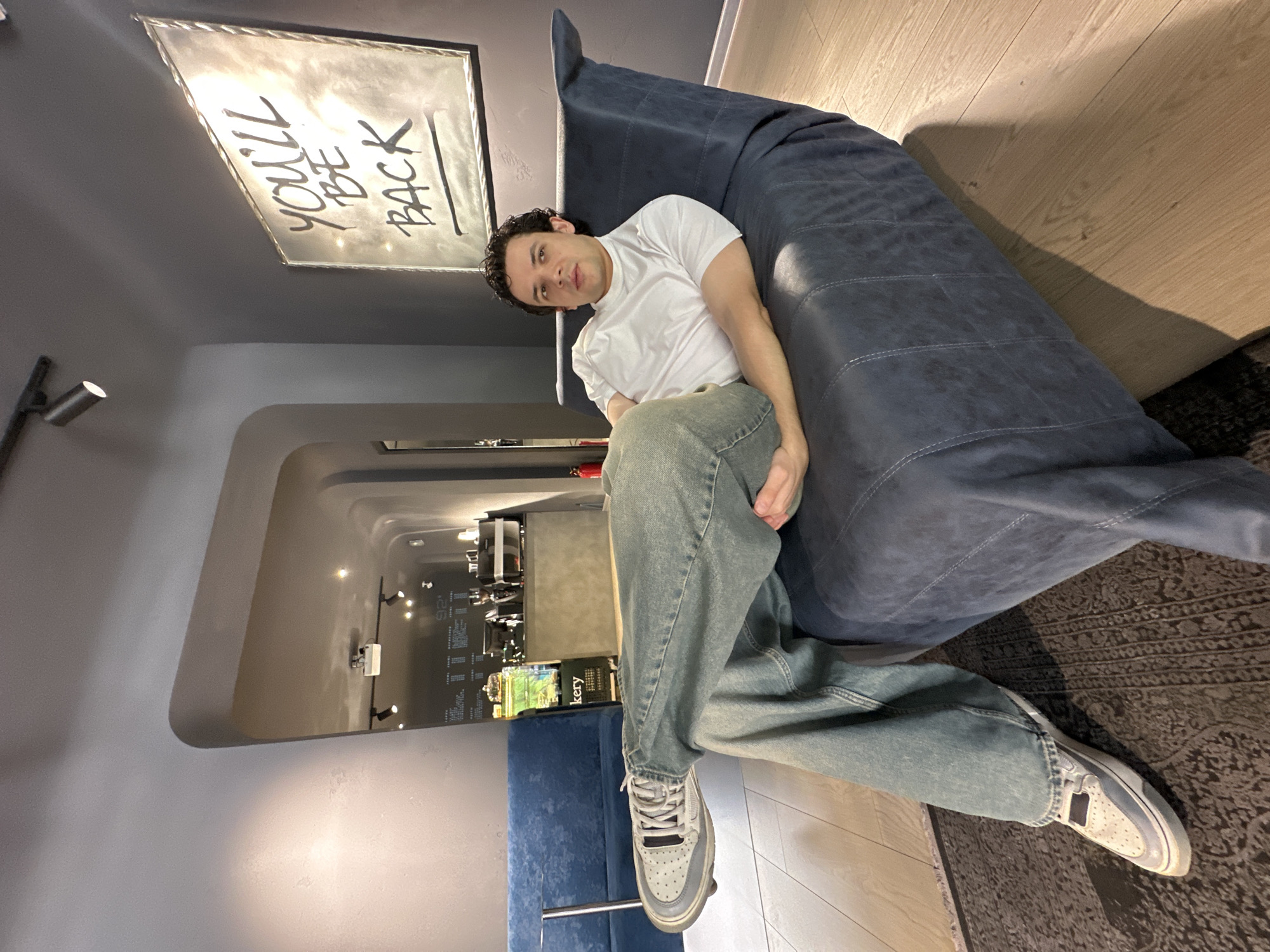 A young man is captured in a modern indoor setting, likely a cafe or lounge in Tashkent, Uzbekistan, sitting on a large, dark blue upholstered sofa or beanbag. He is positioned angled towards the camera, leaning back with a neutral expression, wearing a white t-shirt, light-wash wide-leg jeans, and grey and white sneakers. His dark, curly hair is visible.
Behind and above him, on a dark grey wall, a rectangular framed artwork is prominently displayed, illuminated from an unseen light source. The artwork features bold, handwritten text that reads: "YOU'LL BE BACK".
The flooring consists of light-colored wooden planks transitioning into a dark, patterned rug beneath the seating area. The background reveals distinct architectural features, including smoothly curved walls and ceilings that create a corridor-like depth, illuminated by several small spotlights. Further in the distance, through an opening, a service area or counter is visible with some reflective surfaces and faint text, including "bakery" and "92" (possibly a number or part of a display). Another blue seating element is partially visible in the far background. The overall lighting is artificial, contributing to the contemporary ambiance of the space.