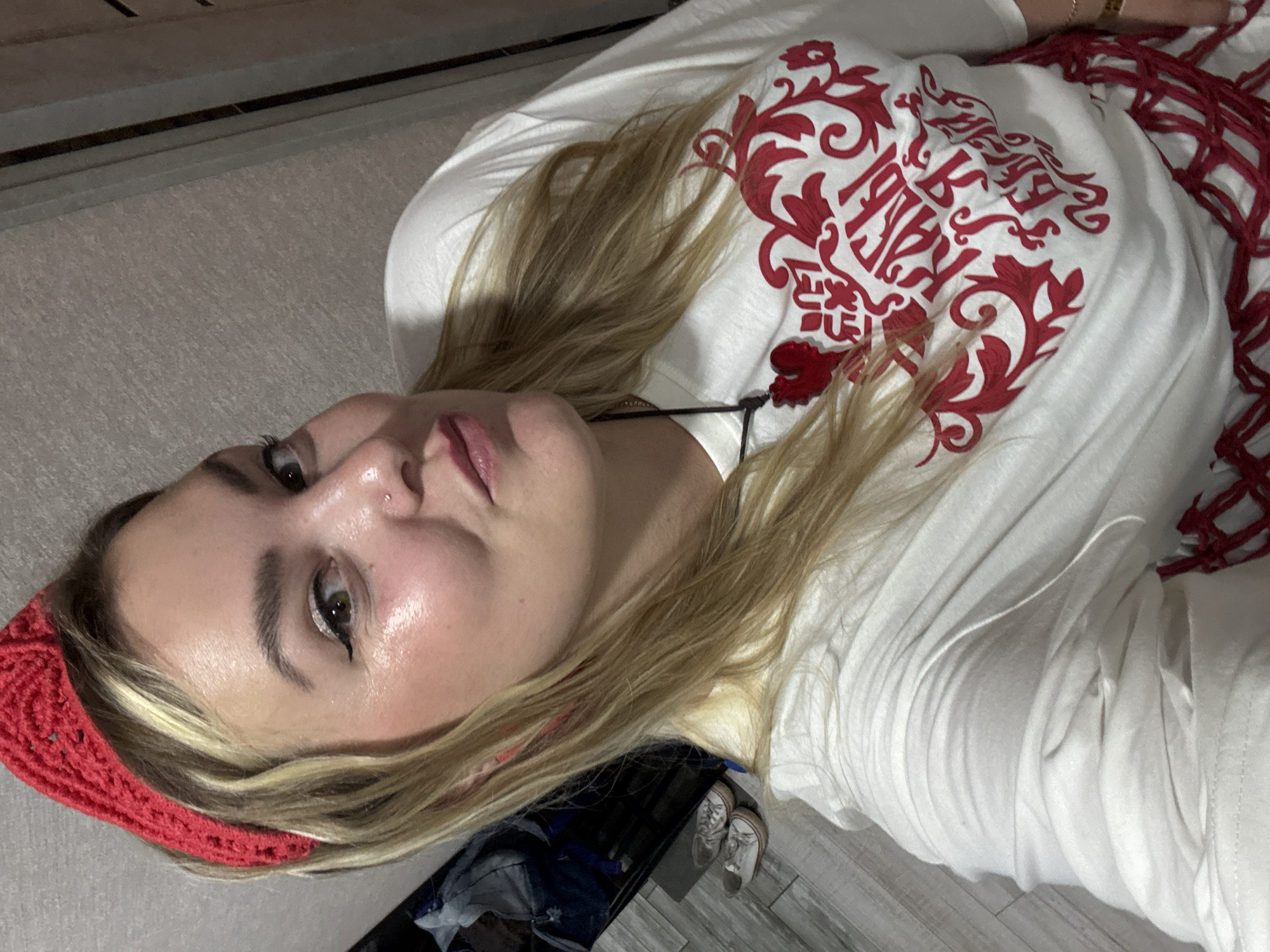 A young woman with blonde hair and light skin is taking a selfie. She is wearing a red knitted headband, a white long-sleeved t-shirt with red ornate lettering on the front, and a red pendant on a black cord. She has winged eyeliner and a nose ring. In the background, there is a wall with a textured pattern, and to the left, a floor with wooden planks and a pair of white sneakers. The scene appears to be indoors.