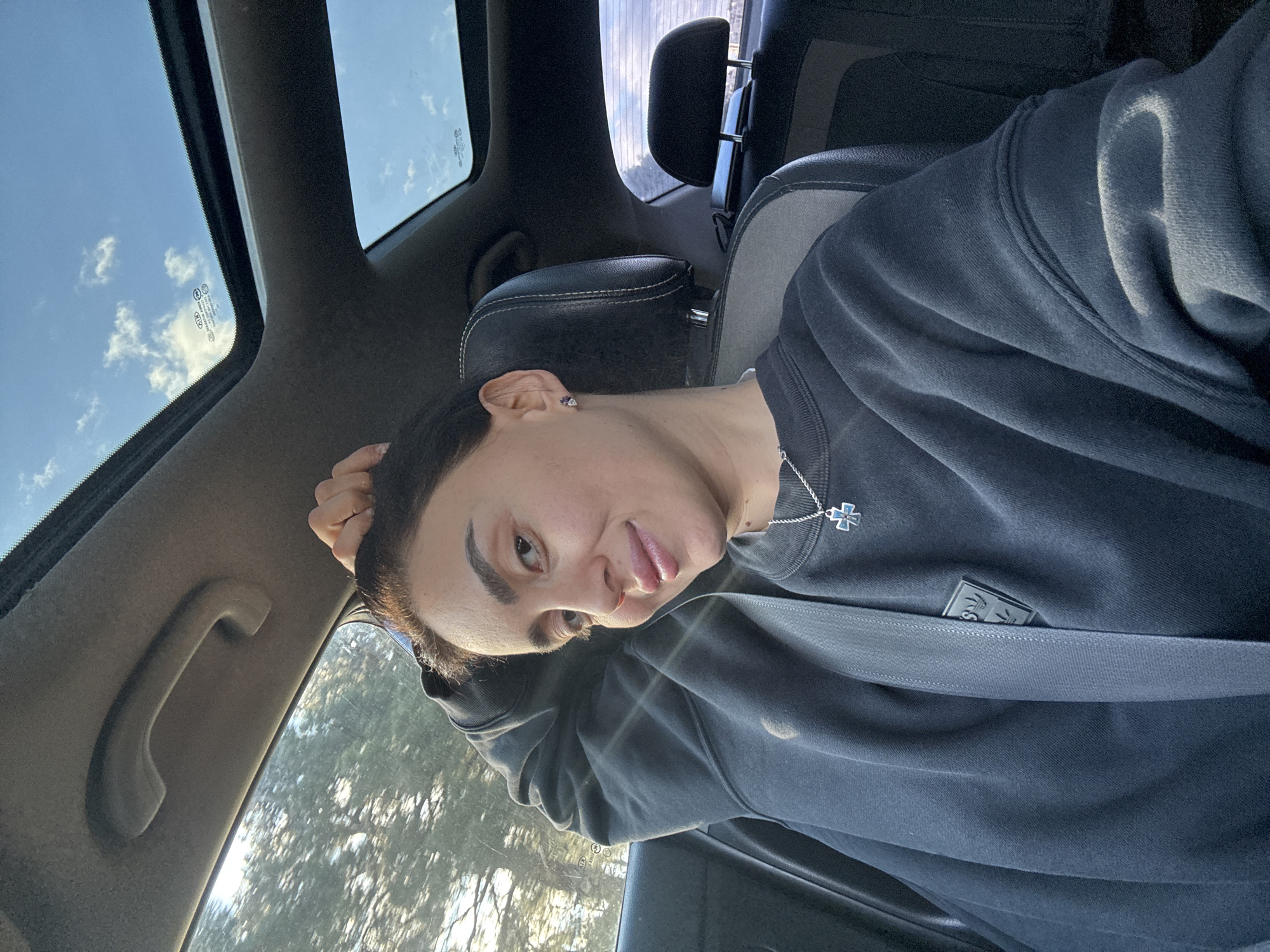 A person is seated inside a vehicle, likely a car, wearing a dark-colored sweatshirt with a seatbelt fastened across their chest. The person has short, dark hair and is wearing a silver necklace with a pendant. The vehicle has a sunroof, through which a blue sky with clouds can be seen. The interior of the car is primarily dark gray and black. Part of the exterior is visible through the side window, showing foliage.
It is not possible to determine the location as Tbilisi, Georgia, from the image alone.