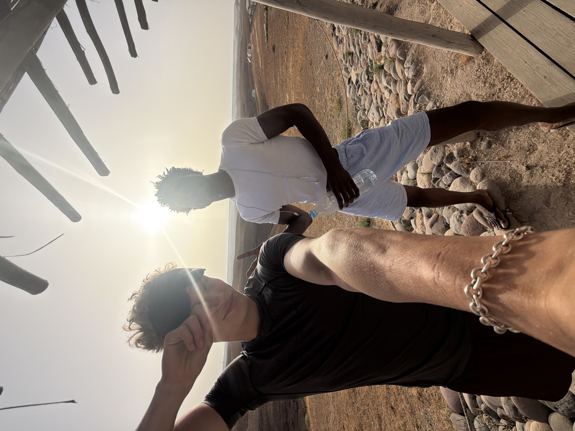 In Agafay, Morocco, two young men are captured in a candid outdoor photograph under a bright, hazy sun. The foreground features a hand wearing a silver chain bracelet, holding the camera, suggesting a selfie. To the left, a young man with blonde, curly hair, wearing a dark t-shirt and a black headband, shields his eyes with his arm while looking at the camera with a slight smile. His gesture implies he is protecting himself from the sun's glare. To the right, a young man with dark, curly hair, dressed in a white t-shirt and light blue shorts, holds a clear plastic bottle of water in his left hand. He is facing forward, with his head angled slightly upwards, also appearing to be affected by the sun's brightness.
The background depicts a dry, arid landscape characteristic of Agafay, with rolling hills covered in sparse brown vegetation. In the distance, several white structures, possibly tents or small buildings, are visible, hinting at a settlement or tourist camp. The ground beneath the men is a mixture of earth and numerous rounded, light-colored stones. Above them, a wooden structure with horizontal beams is partially visible, providing some overhead shade. The overall scene conveys a sense of casual outdoor activity in a desert environment.