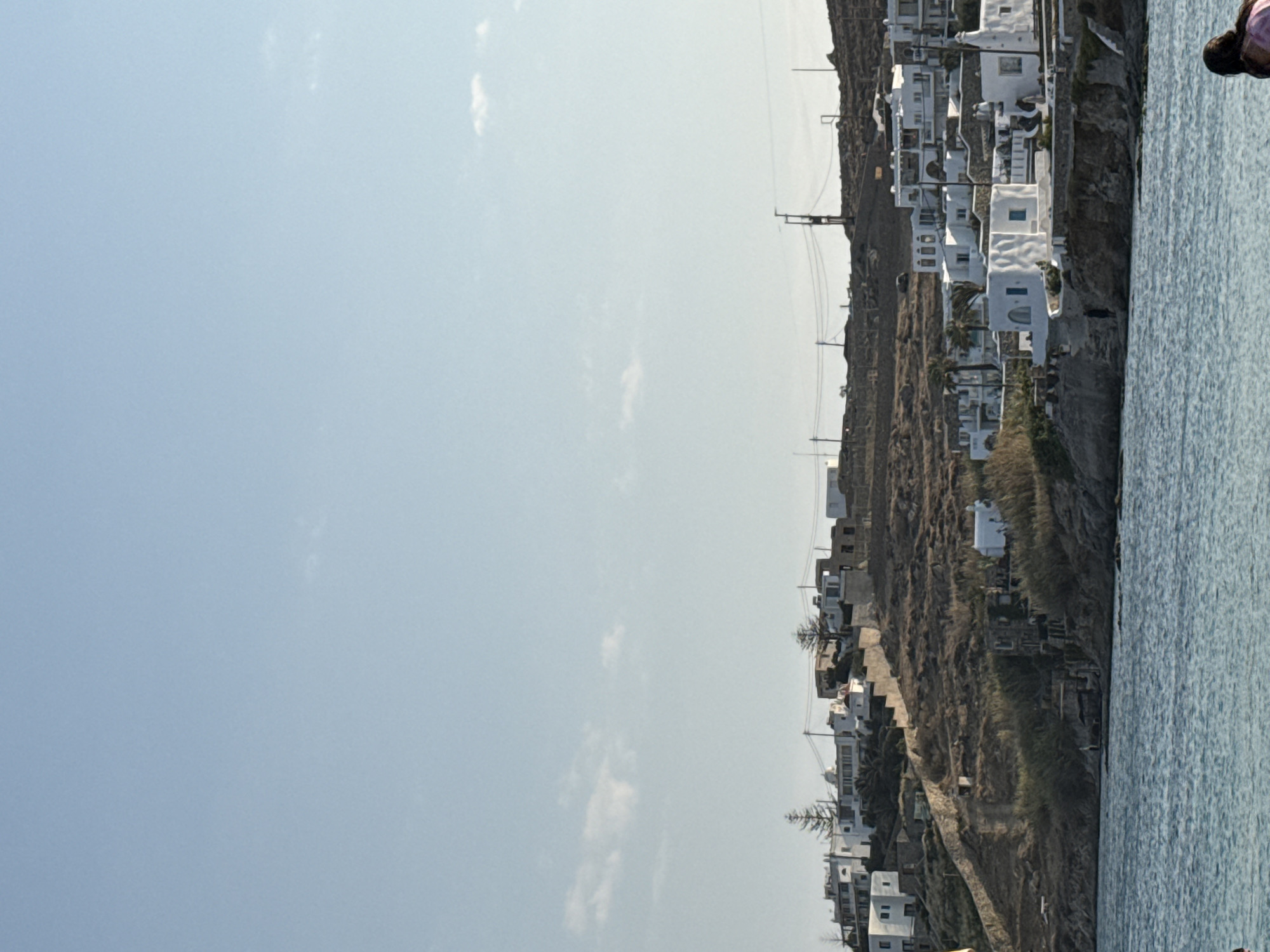 In the city of Agios Ioannis Diakoftis, Greece, the image depicts a coastal scene with a bright, clear blue sky and a few wispy clouds. The foreground is dominated by the turquoise waters of the sea. On the right side of the frame, a person with dark hair, wearing a pink top, is partially visible, facing away from the camera. The background features a hillside covered in dry vegetation and rocky terrain. Dotted along the hillside are numerous whitewashed buildings with flat roofs, characteristic of Mediterranean architecture. Some of these buildings are clustered together, forming a small village or settlement, while others appear more isolated. There are also visible power lines and utility poles running across the hillside. The overall impression is one of a sun-drenched, tranquil coastal environment.